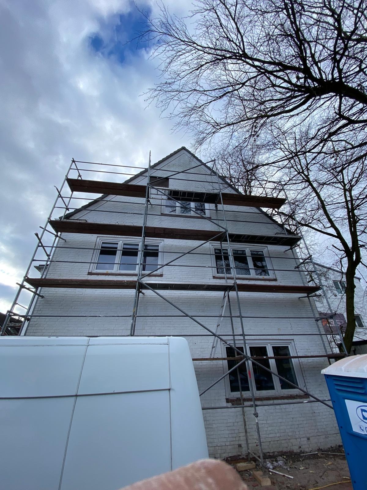 Biały dom w rusztowaniach, widok z dołu, częściowo zasłonięty przez biały samochód dostawczy. Drzewo z nagimi gałęziami po prawej stronie.