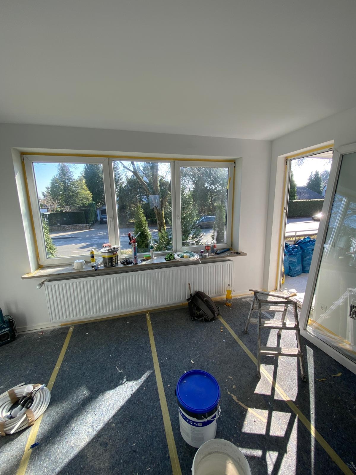 Pokój w trakcie malowania: zabezpieczone okna i podłoga, farby i narzędzia na parapecie. Widok na zewnątrz przez okno. Przygotowania do malowania ścian.