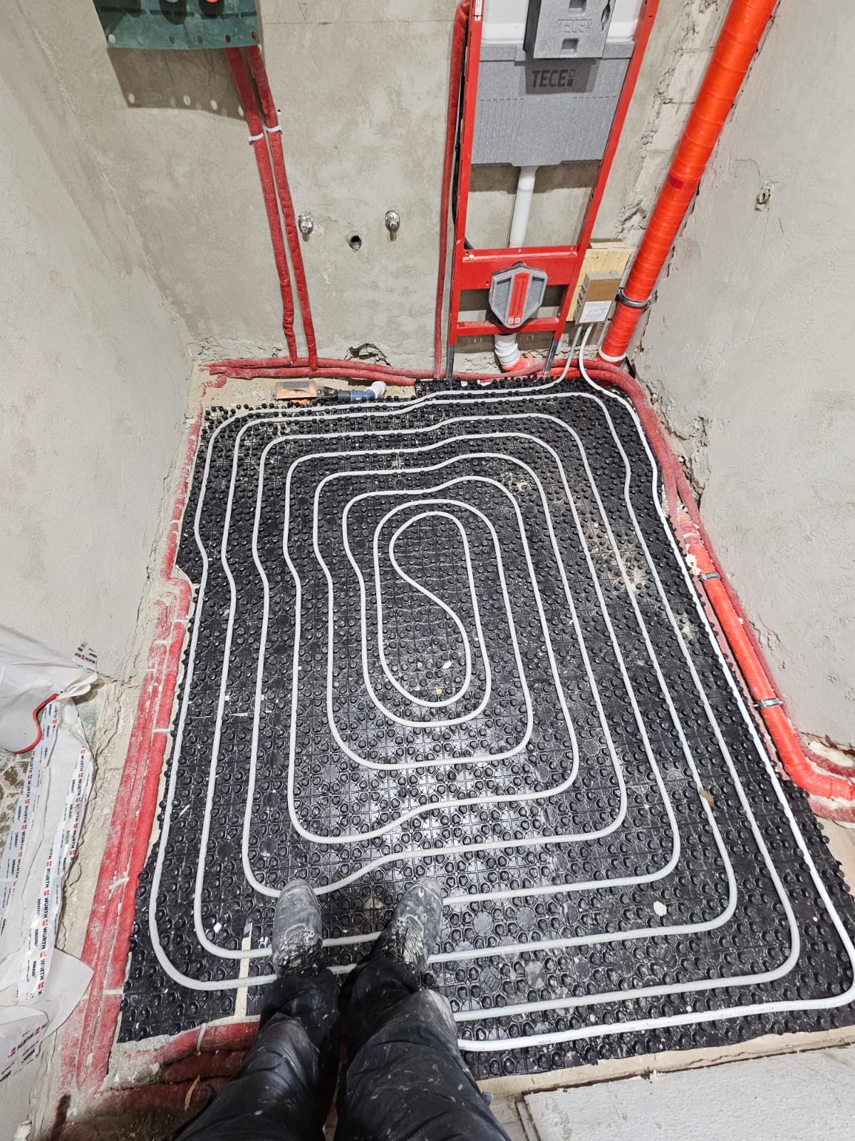 Ułożona instalacja ogrzewania podłogowego z widocznymi rurami w kształcie spirali, przed wylaniem posadzki, z elementami instalacji sanitarnej na ścianach.