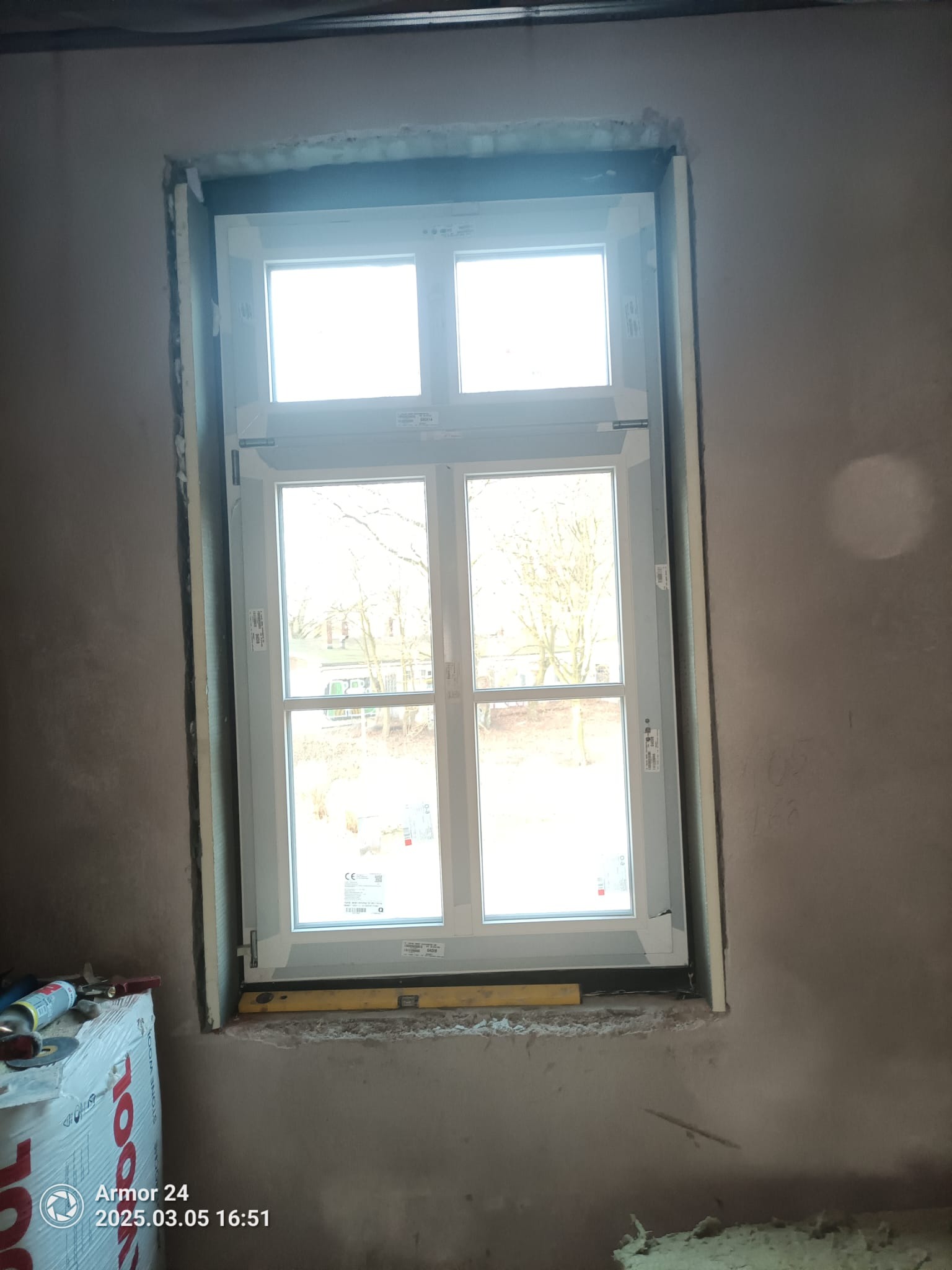 Białe okno PCV zamontowane w surowej wnęce, z widocznymi narzędziami i materiałami montażowymi. W tle widok na zewnątrz.