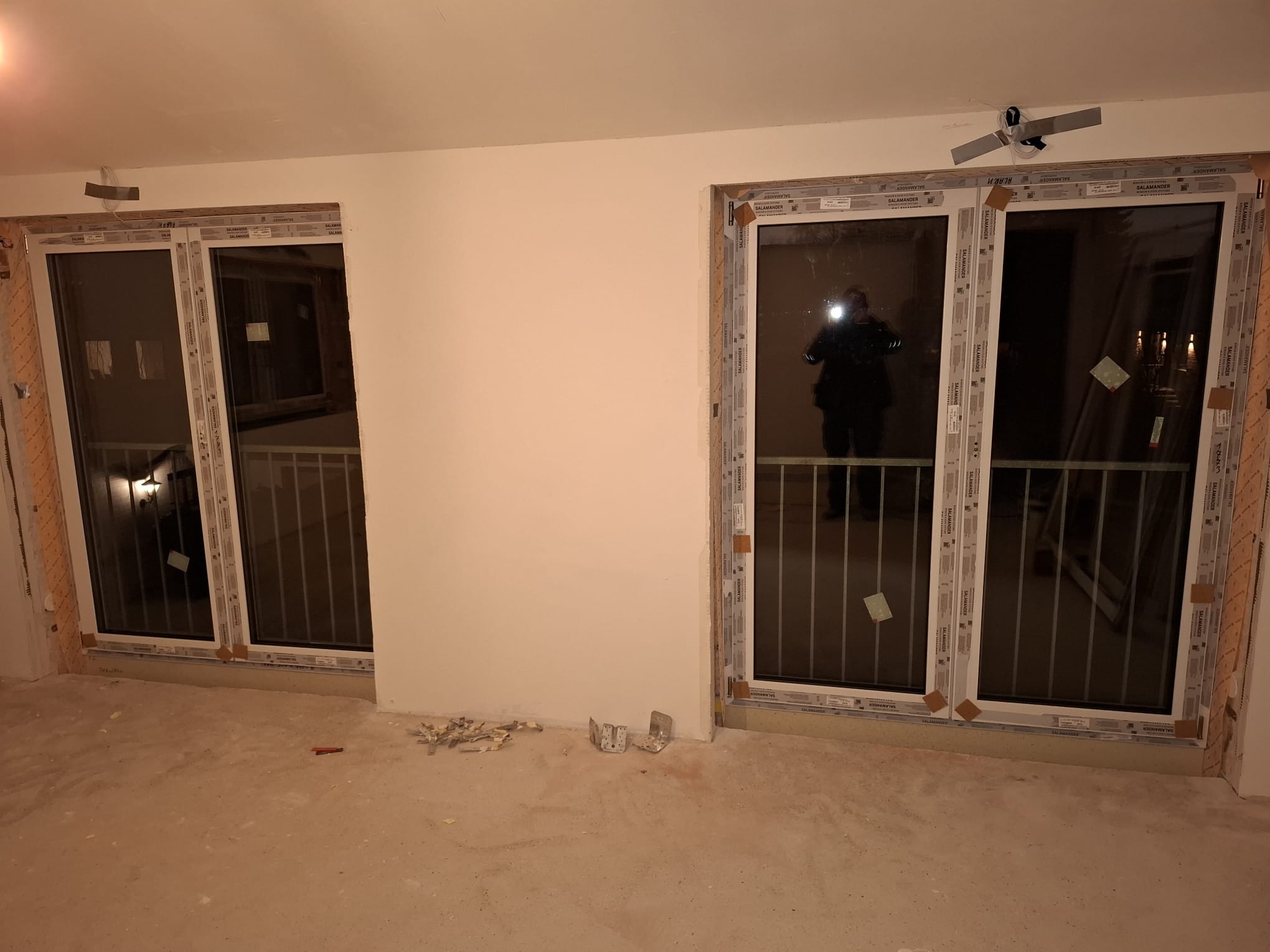 Montaż okien PCV Salamander: widok na balkon przez nowe okna z białymi ramami, w trakcie prac wykończeniowych, z widocznym odbiciem osoby w szybie.