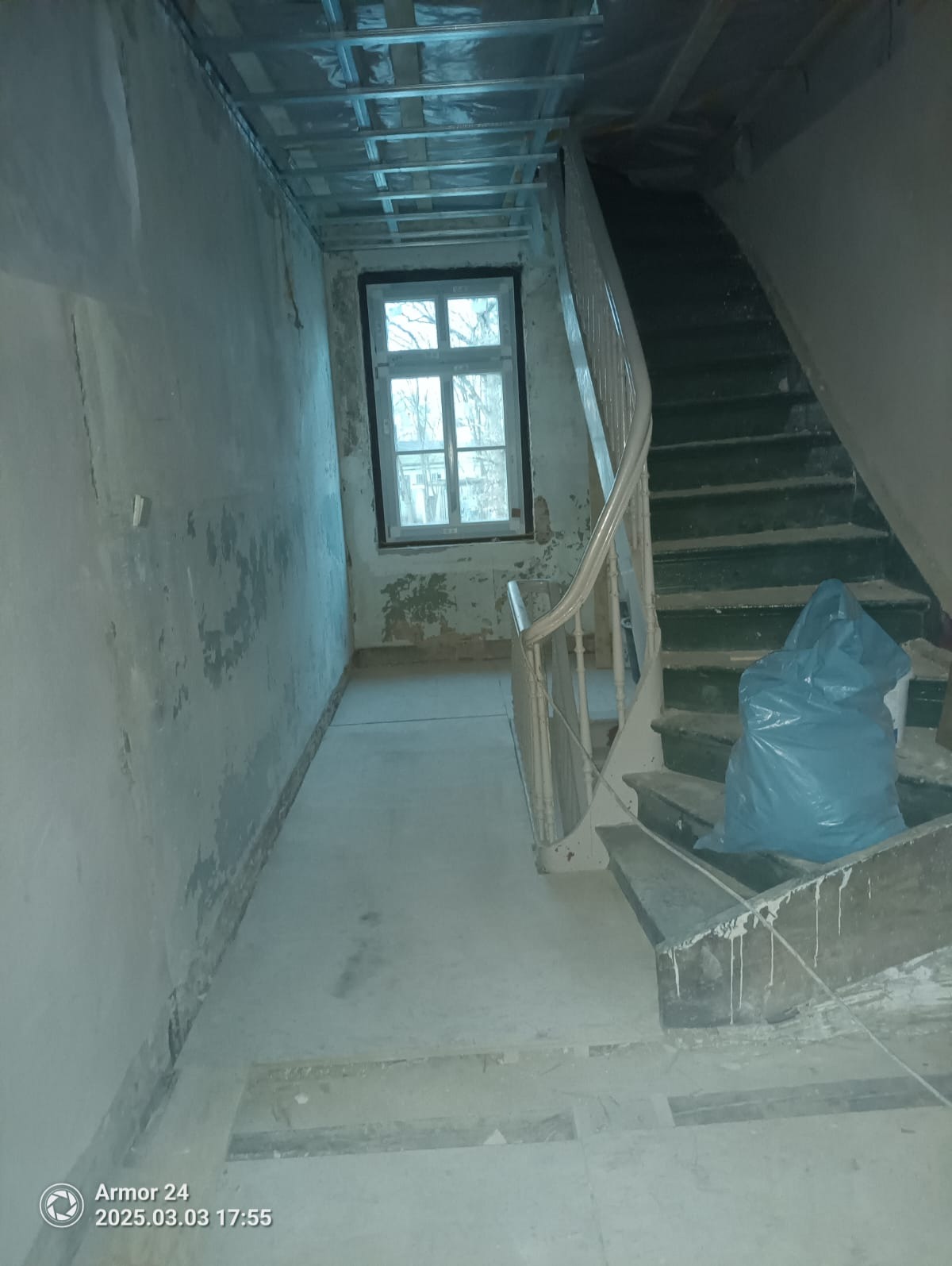 Wnętrze w trakcie remontu: surowe ściany z odpadającym tynkiem, okno z widokiem na drzewa, schody z workiem gruzu i metalowa konstrukcja sufitu podwieszanego.