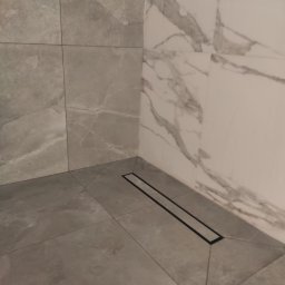 Lucas Serwis Budowlany - Wykończony narożnik kabiny prysznicowej z odpływem liniowym. Szare płytki podłogowe i marmurowe na ścianach. Minimalistyczny design.