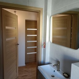 Lucas Serwis Budowlany - Nowoczesna łazienka z drewnianymi drzwiami z przeszkleniami, umywalką nablatową i lustrem. Widoczne płytki w odcieniach szarości i brązowe płytki podłogowe.