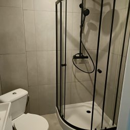 Lucas Serwis Budowlany - Nowoczesna kabina prysznicowa z czarnymi profilami w łazience wykończonej szarymi płytkami. Widoczna toaleta i umywalka. Minimalistyczny design.