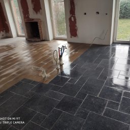 Lucas Serwis Budowlany - Układanie ciemnych płytek podłogowych w pomieszczeniu z przygotowanym podłożem. Widoczne narzędzia glazurnicze i częściowo wykończone ściany.