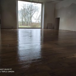 Lucas Serwis Budowlany - Ułożona, błyszcząca podłoga z paneli w surowym wnętrzu z dużym oknem na zewnątrz. Widoczne niedokończone ściany i sufit. Minimalistyczny, nowoczesny styl.