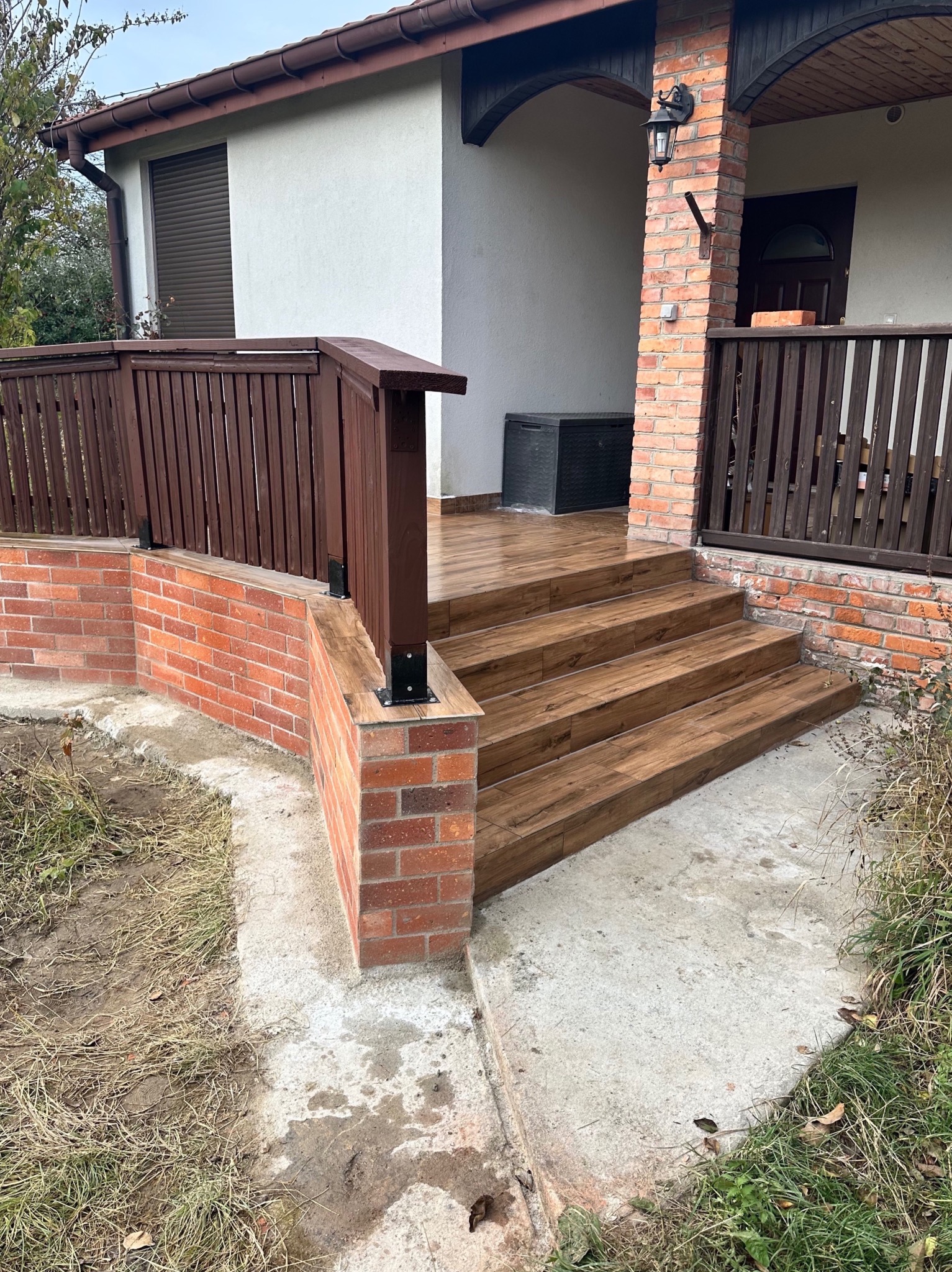 Wykończone schody wejściowe z brązowych płytek imitujących drewno, obłożone cegłą, z drewnianą balustradą. Widok na elewację domu z białym tynkiem i brązowymi elementami.