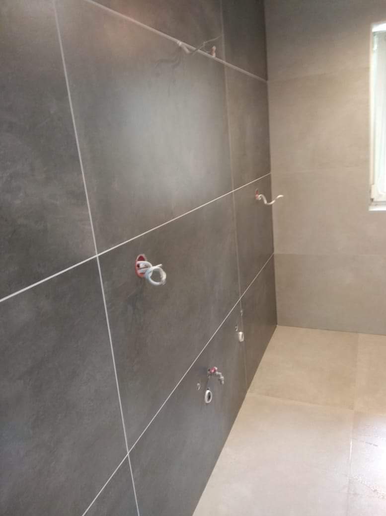 Ciemnoszare płytki ścienne z białymi fugami i wystającymi elementami instalacji wodnej w łazience, widok z perspektywy.