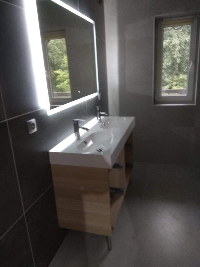 Nowoczesna łazienka z podwójną umywalką na drewnianej szafce, podświetlanym lustrem i oknem z widokiem na zieleń. Szare płytki na ścianach i podłodze.