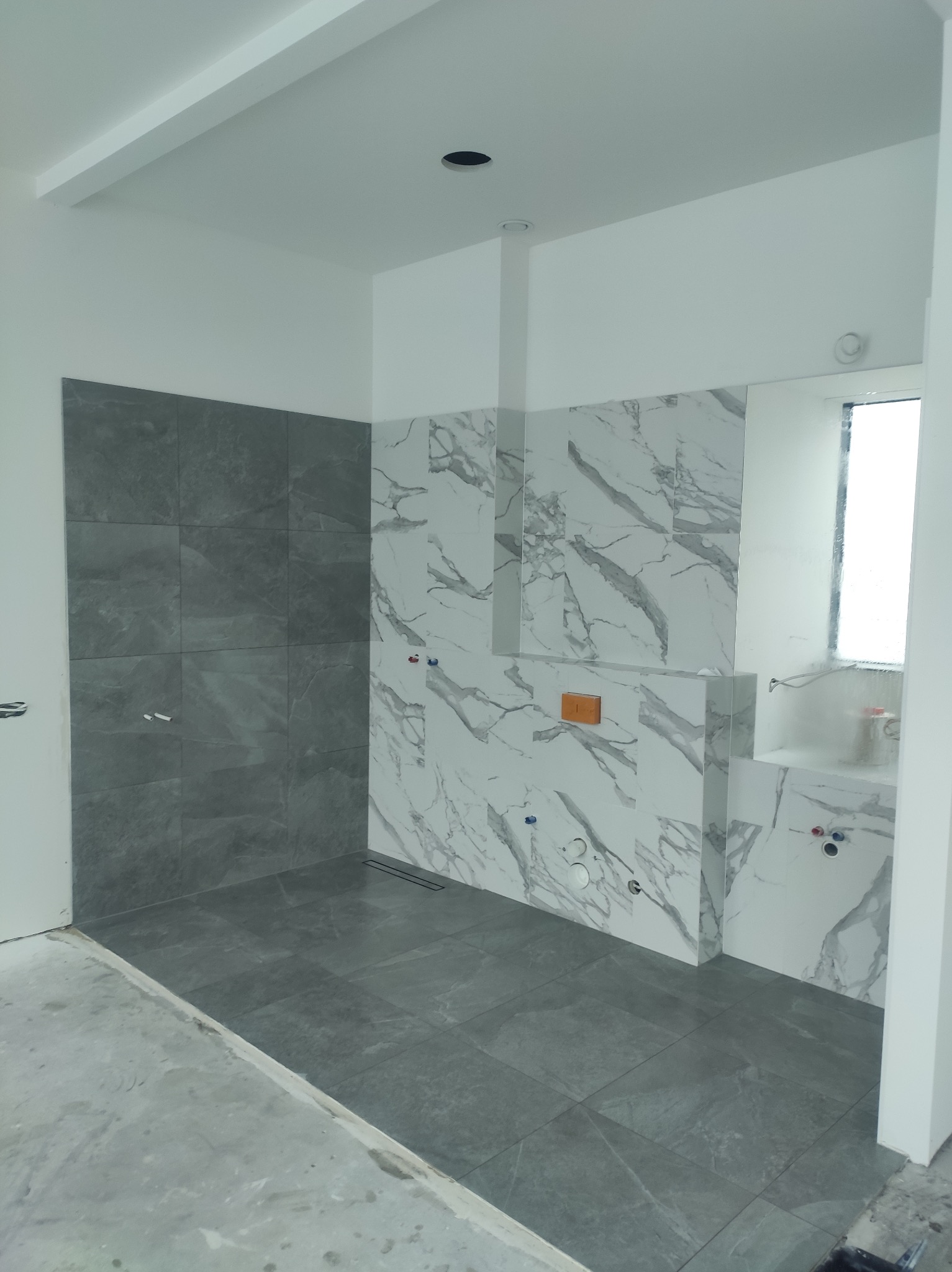 Wykończona łazienka z szarymi i marmurkowymi płytkami, widoczne podejścia hydrauliczne i odpływ liniowy. Podłoga w szarych płytkach. Stan surowy.