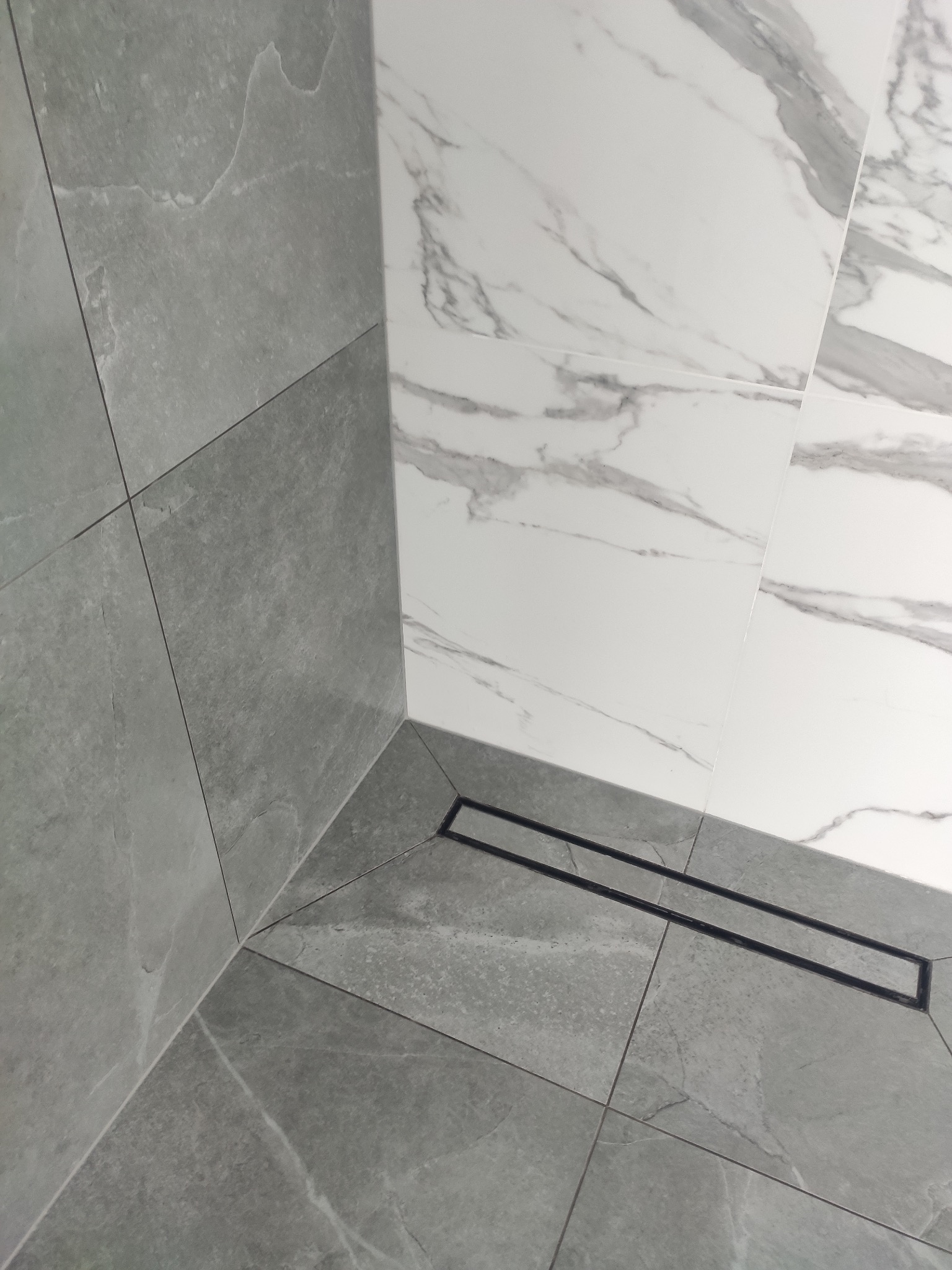 Nowoczesna łazienka z szarymi i białymi płytkami imitującymi marmur oraz czarnym odpływem liniowym w rogu kabiny prysznicowej.