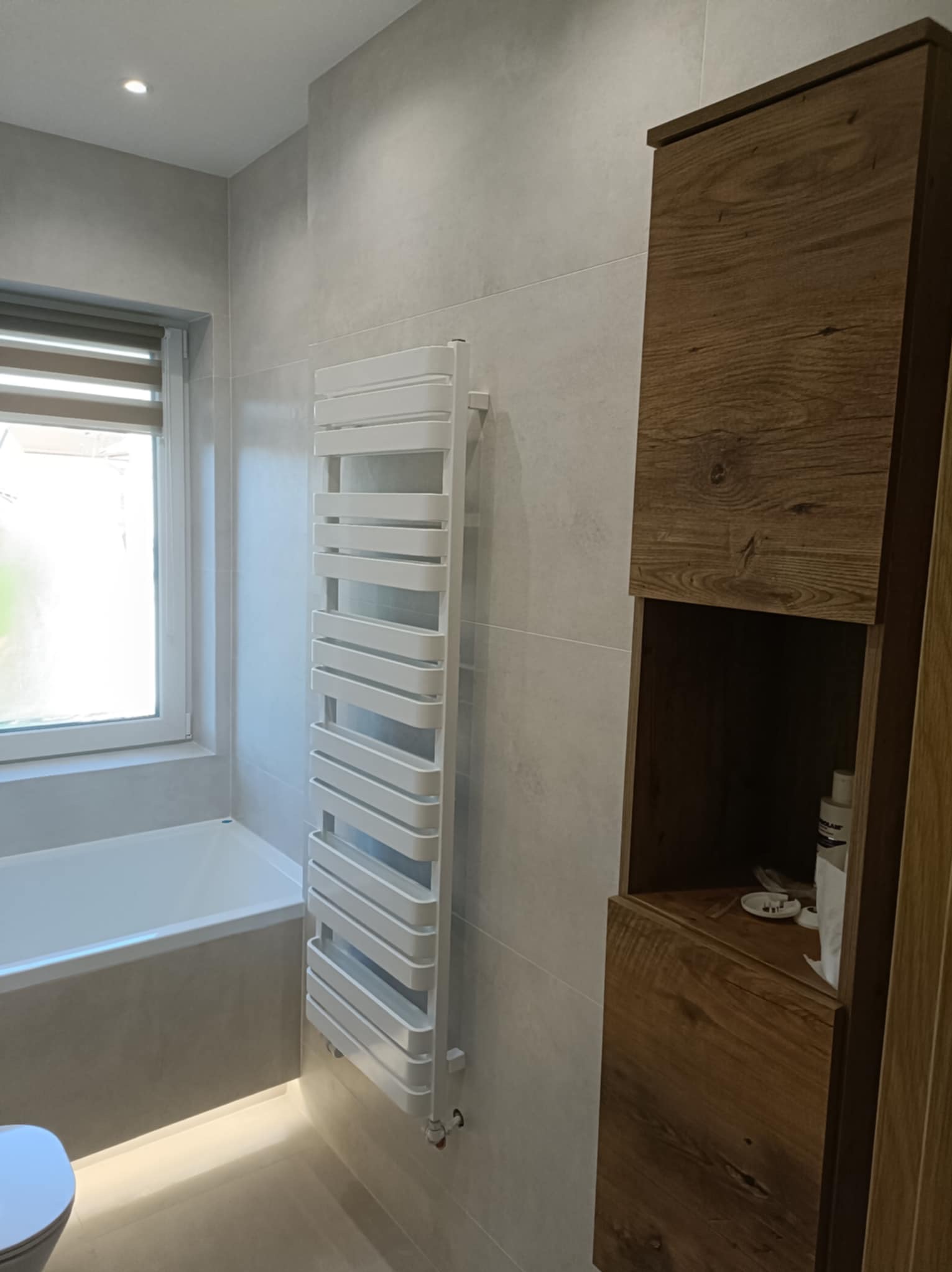 Nowoczesna łazienka w szarościach z drewnianą szafką, grzejnikiem drabinkowym i oknem. Minimalistyczny design z subtelnym oświetleniem.