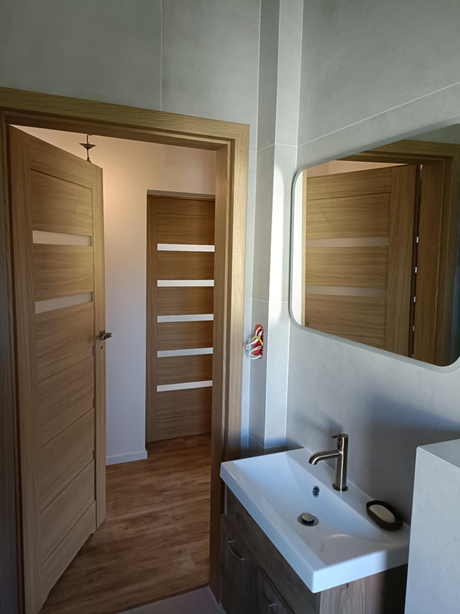 Nowoczesna łazienka z drewnianymi drzwiami z przeszkleniami, umywalką nablatową i lustrem. Widoczne płytki w odcieniach szarości i brązowe płytki podłogowe.