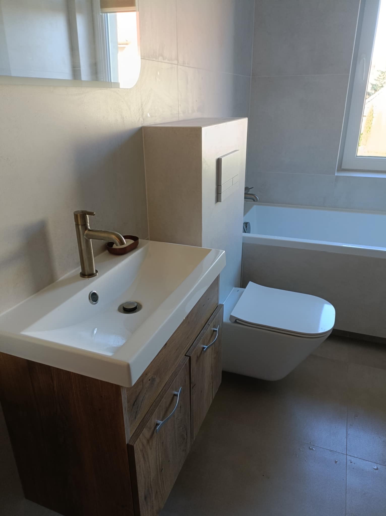 Nowoczesna łazienka z umywalką na drewnianej szafce, podwieszaną toaletą i wanną. Jasne płytki na ścianach i podłodze. Minimalistyczny design.