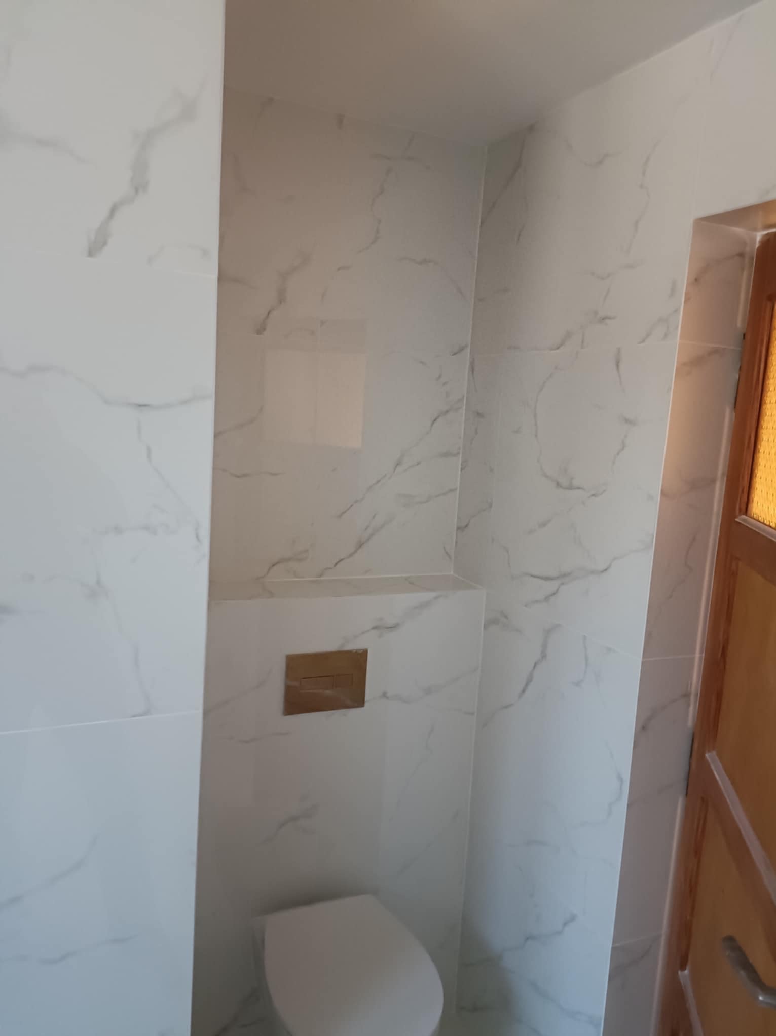 Wykończona łazienka z białymi płytkami imitującymi marmur, zabudowana toaleta z złotym przyciskiem, widoczny fragment drewnianych drzwi. Minimalistyczny design.