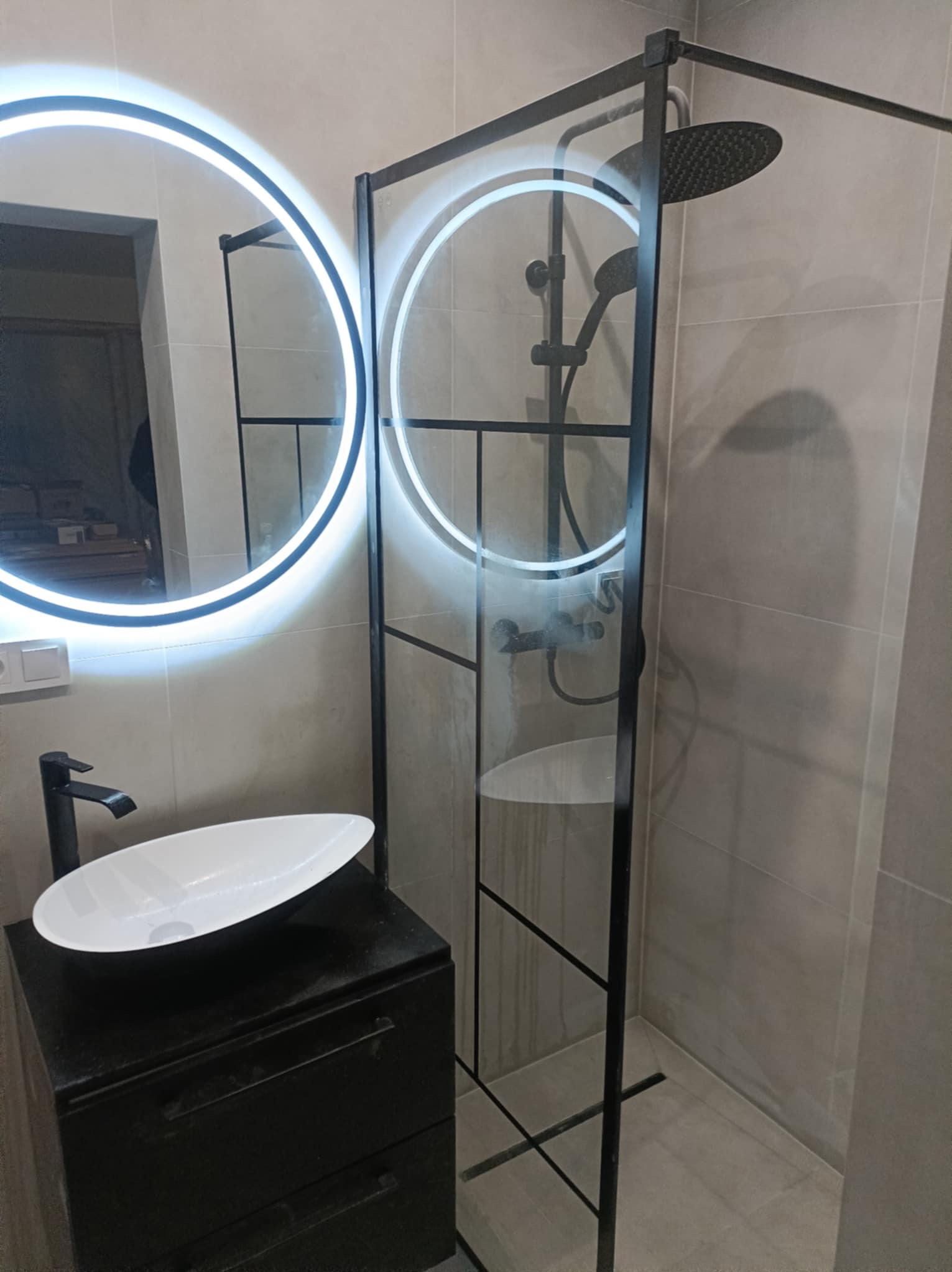 Nowoczesna łazienka z kabiną prysznicową typu walk-in z czarnymi profilami, okrągłym lustrem z podświetleniem LED i umywalką nablatową na czarnej szafce.