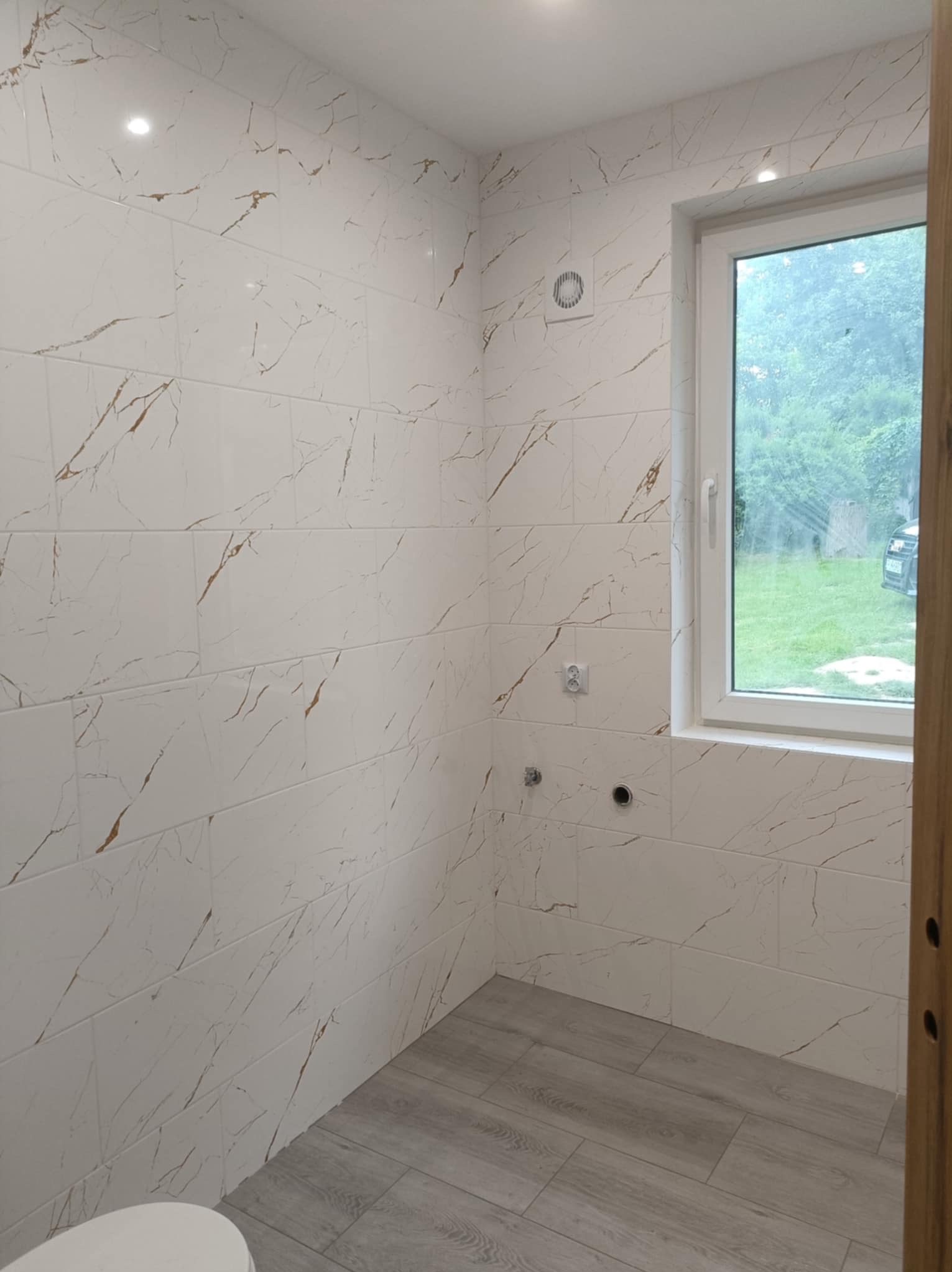 Wykończona łazienka z białymi płytkami imitującymi marmur ze złotymi żyłami, szarą podłogą imitującą drewno i oknem z widokiem na zieleń.