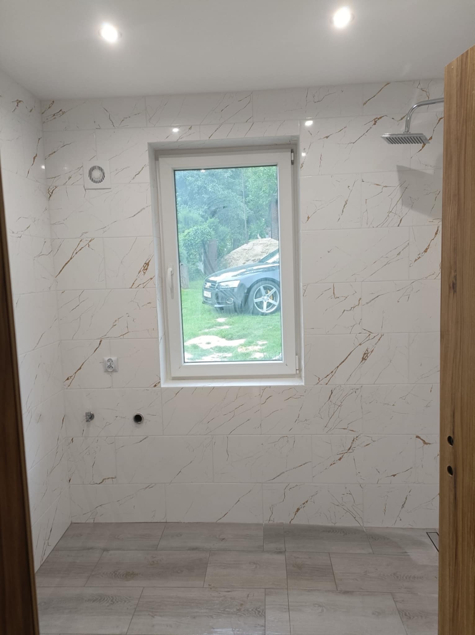 Wykończona łazienka z białymi płytkami imitującymi marmur, widok na okno z samochodem na zewnątrz. Nowoczesna kabina prysznicowa i oświetlenie sufitowe.