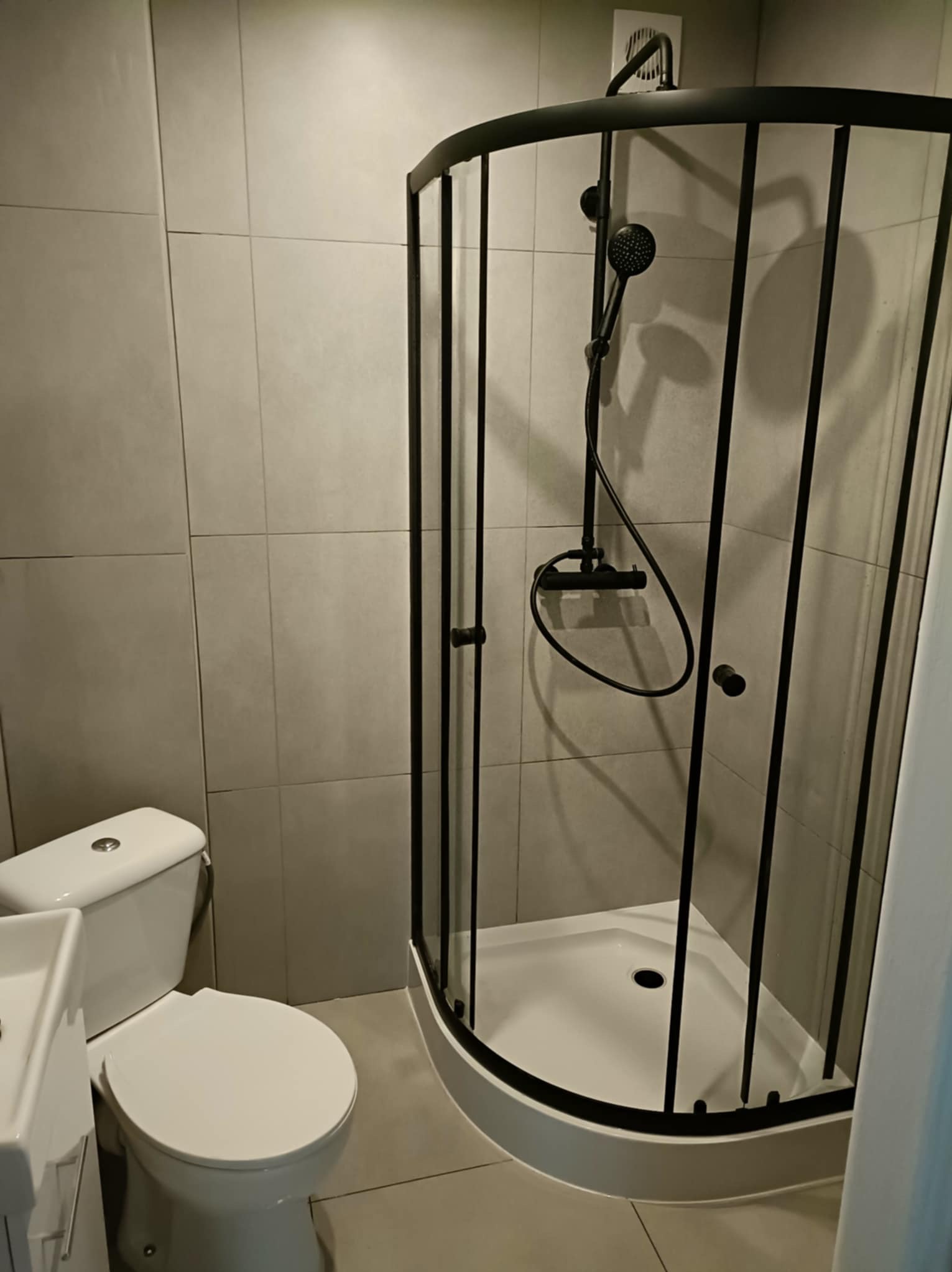 Nowoczesna kabina prysznicowa z czarnymi profilami w łazience wykończonej szarymi płytkami. Widoczna toaleta i umywalka. Minimalistyczny design.