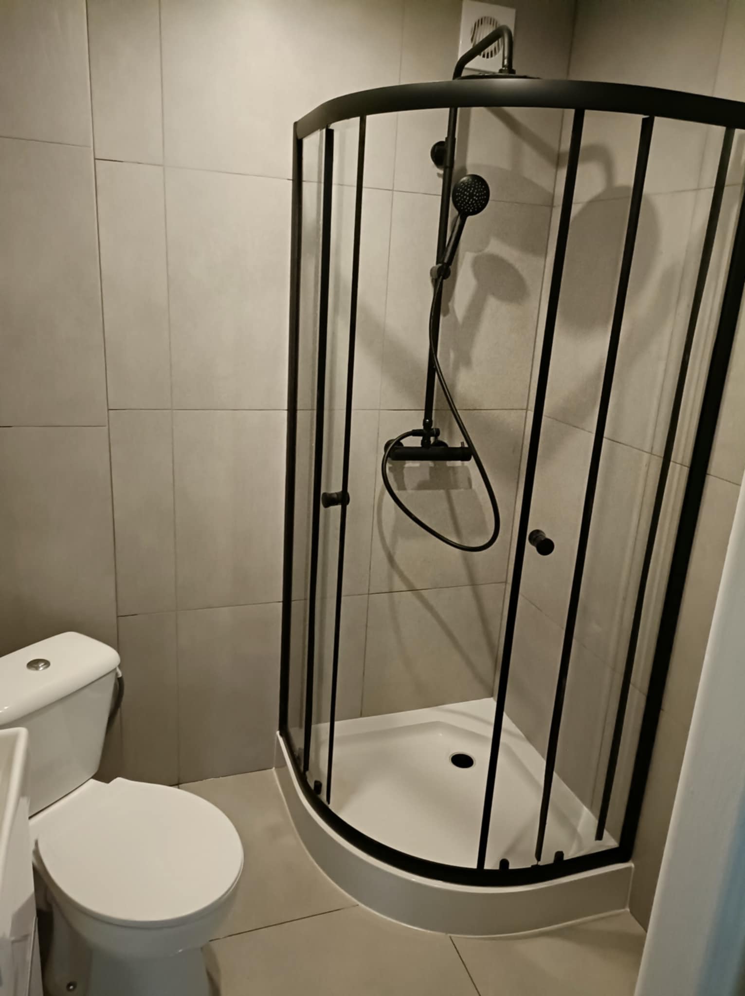 Nowoczesna kabina prysznicowa z czarną ramą i deszczownicą, umieszczona w łazience wykończonej jasnymi, kwadratowymi płytkami. Widoczny fragment toalety.