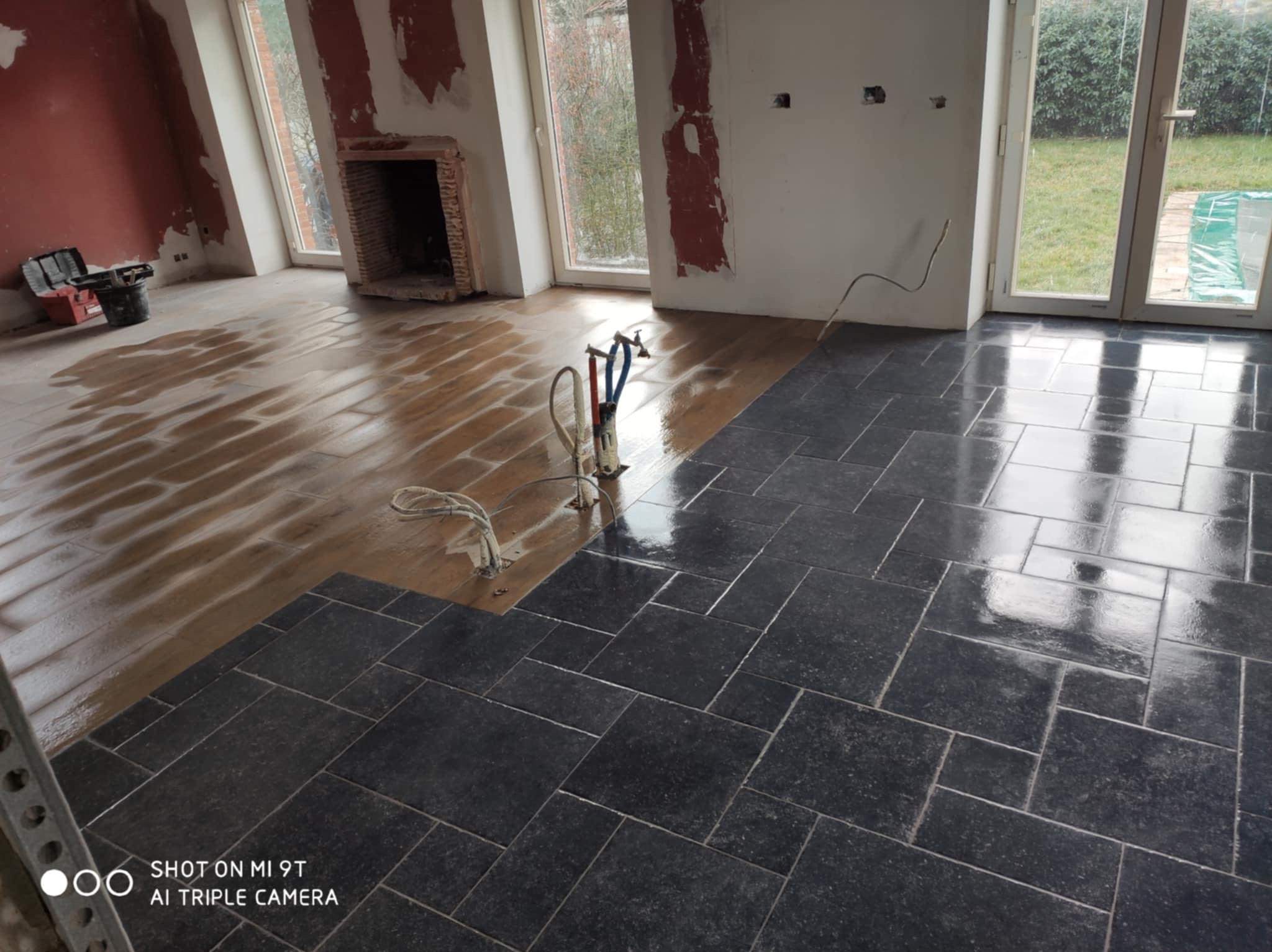 Układanie ciemnych płytek podłogowych w pomieszczeniu z przygotowanym podłożem. Widoczne narzędzia glazurnicze i częściowo wykończone ściany.