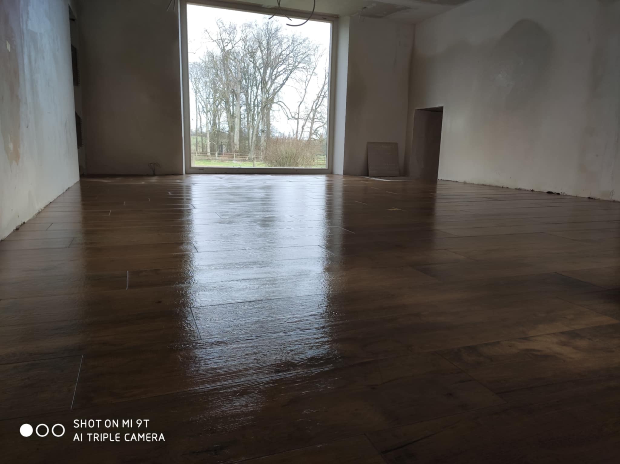 Ułożona, błyszcząca podłoga z paneli w surowym wnętrzu z dużym oknem na zewnątrz. Widoczne niedokończone ściany i sufit. Minimalistyczny, nowoczesny styl.