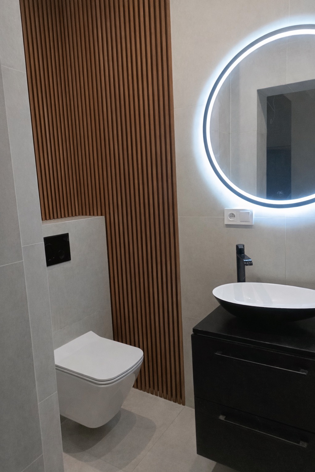 Nowoczesna łazienka z drewnianą ścianką działową, okrągłym lustrem z podświetleniem LED i minimalistyczną umywalką nablatową na czarnej szafce.