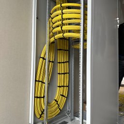 ZID-ELEKTRO PATRYK WASILEWSKI - Żółte kable w metalowej szafie rack, starannie ułożone i zabezpieczone czarnymi opaskami. Widoczne elementy konstrukcyjne szafy i fragment ściany.