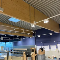 Wnętrze sklepu w trakcie montażu oświetlenia: drewniane belki, białe lampy, sufit z blachy trapezowej, stanowiska kasowe, narzędzia na stole, plany na ścianie.