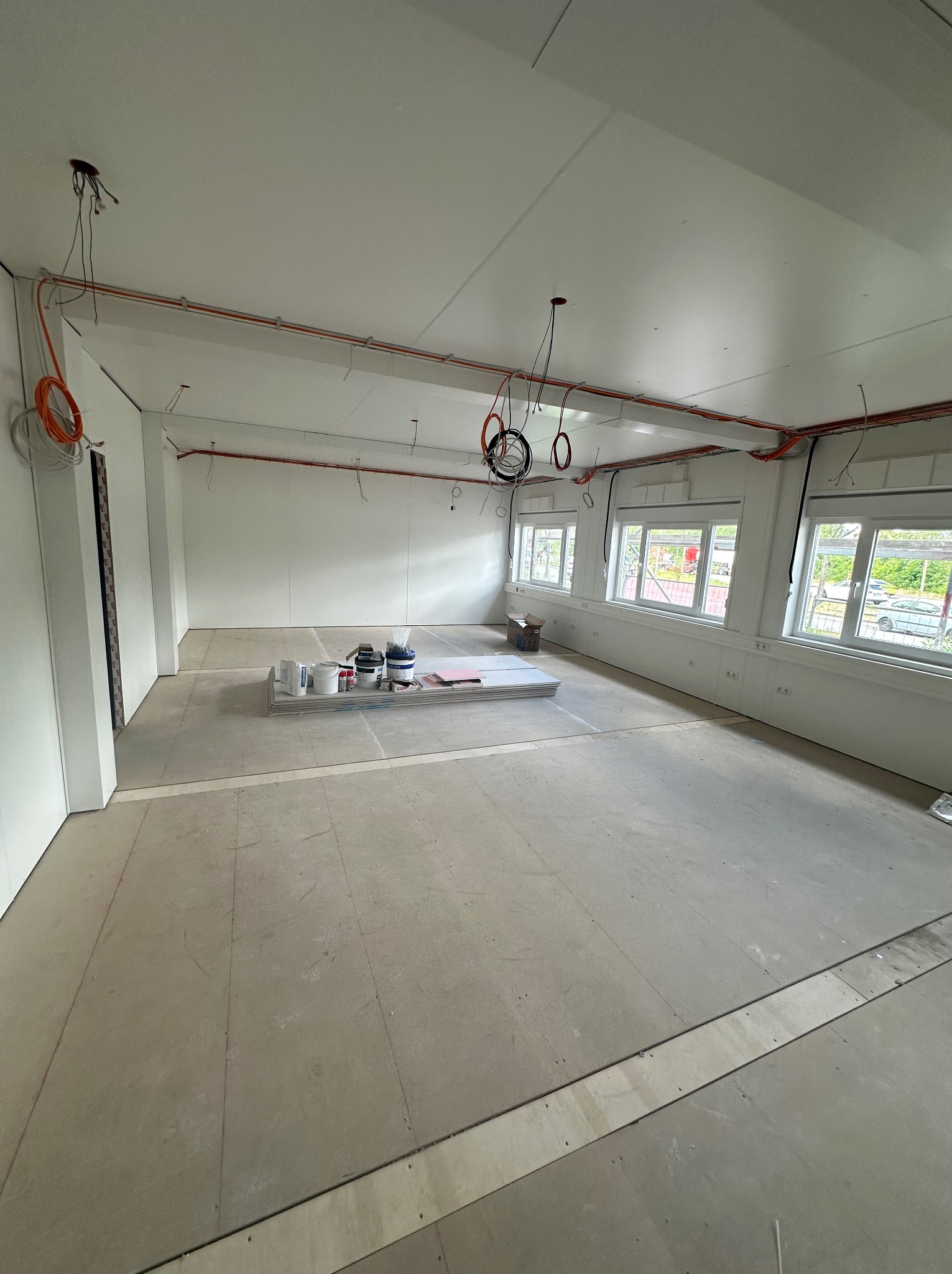 Wnętrze w trakcie wykończenia: widoczne kable elektryczne pod sufitem, ściany przygotowane do malowania, podłoże pod wylewkę. Na podłodze materiały budowlane.