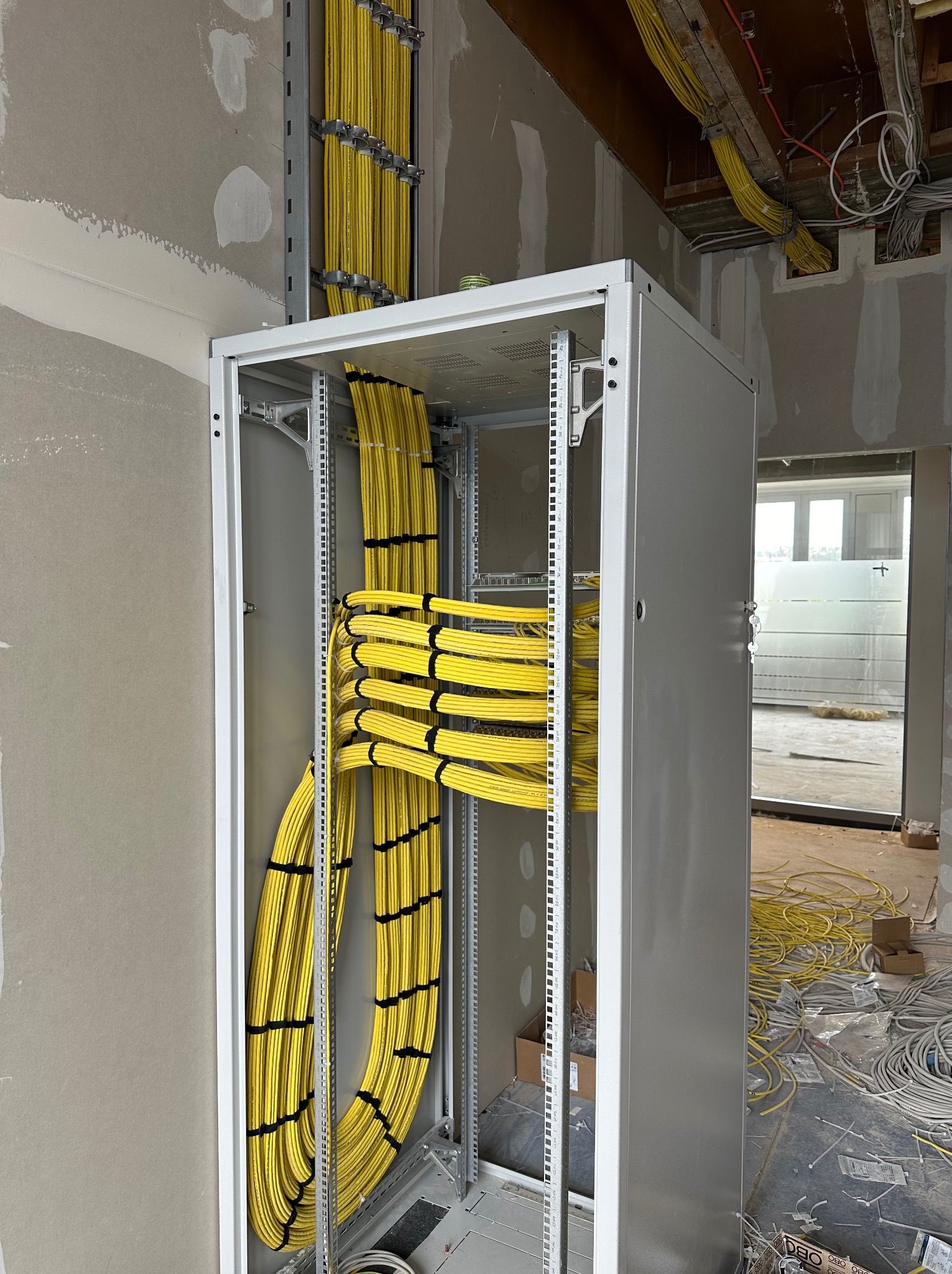 Szafa rack z uporządkowanymi kablami elektrycznymi w żółtej izolacji, zamocowanymi na drabinkach w surowym wnętrzu budynku w trakcie prac wykończeniowych.
