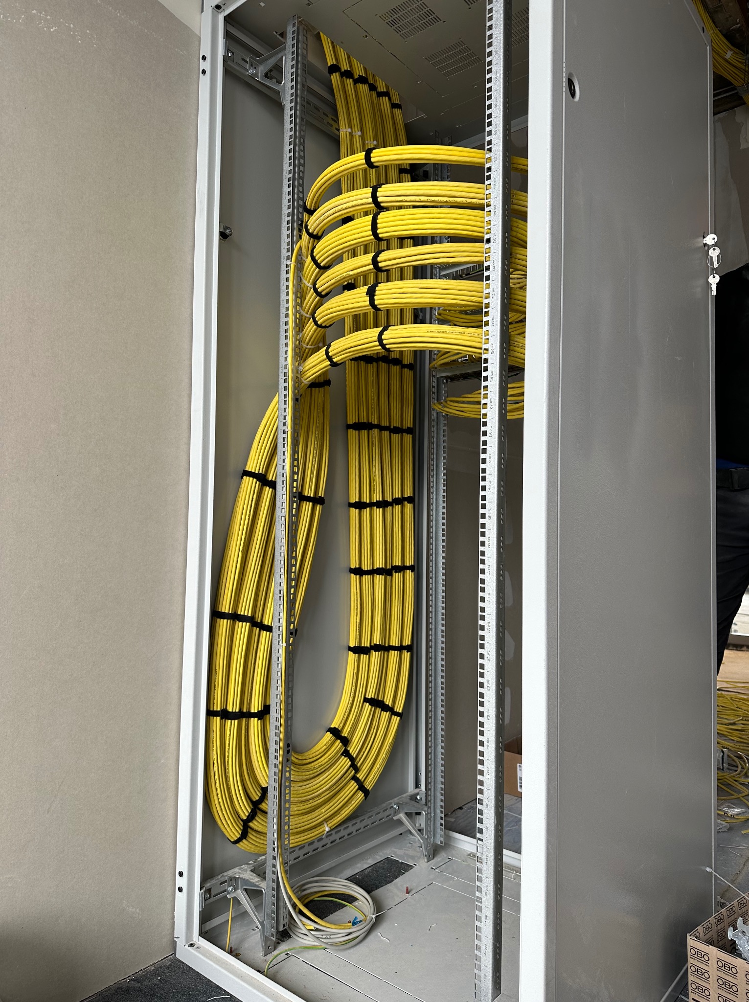 Żółte kable w metalowej szafie rack, starannie ułożone i zabezpieczone czarnymi opaskami. Widoczne elementy konstrukcyjne szafy i fragment ściany.
