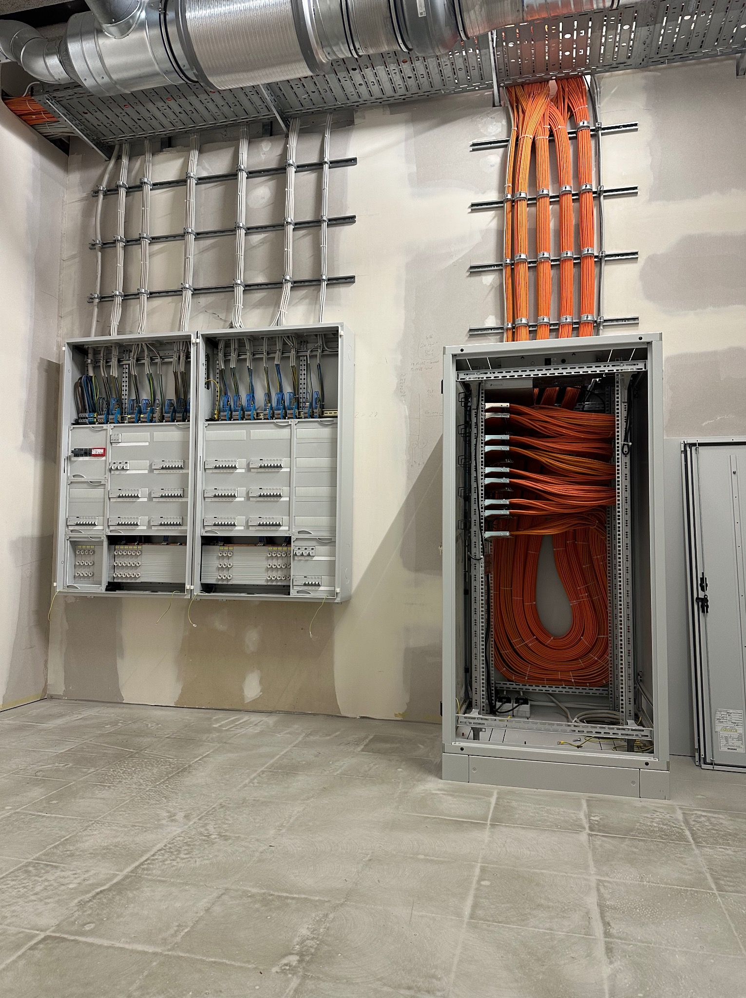 Rozdzielnice elektryczne z okablowaniem w surowym wnętrzu. Widoczne kable białe i pomarańczowe, metalowe koryta kablowe pod sufitem, ściany w trakcie wykończenia.