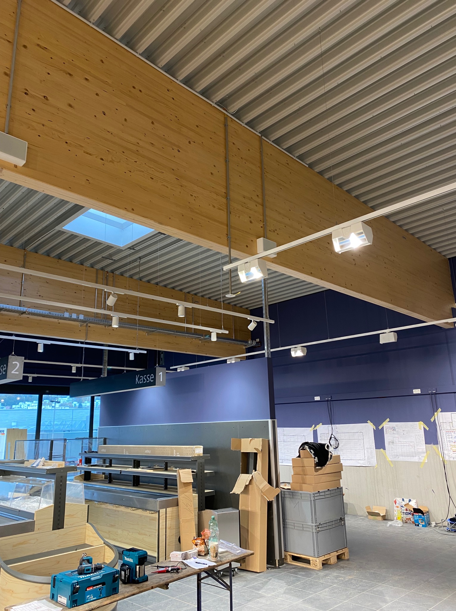 Wnętrze sklepu w trakcie montażu oświetlenia: drewniane belki, białe lampy, sufit z blachy trapezowej, stanowiska kasowe, narzędzia na stole, plany na ścianie.