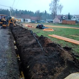 MM INSTAL-WIERT Marcin Twaróg - Prace ziemne: wykop pod rury, żółty mini-koparka w akcji, pomarańczowe rury ułożone na trawie. Domy w tle, pochmurne niebo.