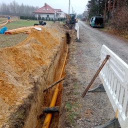 MM INSTAL-WIERT Marcin Twaróg - Żółte rury w wykopie ziemnym, zabezpieczone białą barierką. W tle domy jednorodzinne i sprzęt budowlany. Prace ziemne na etapie układania rur.