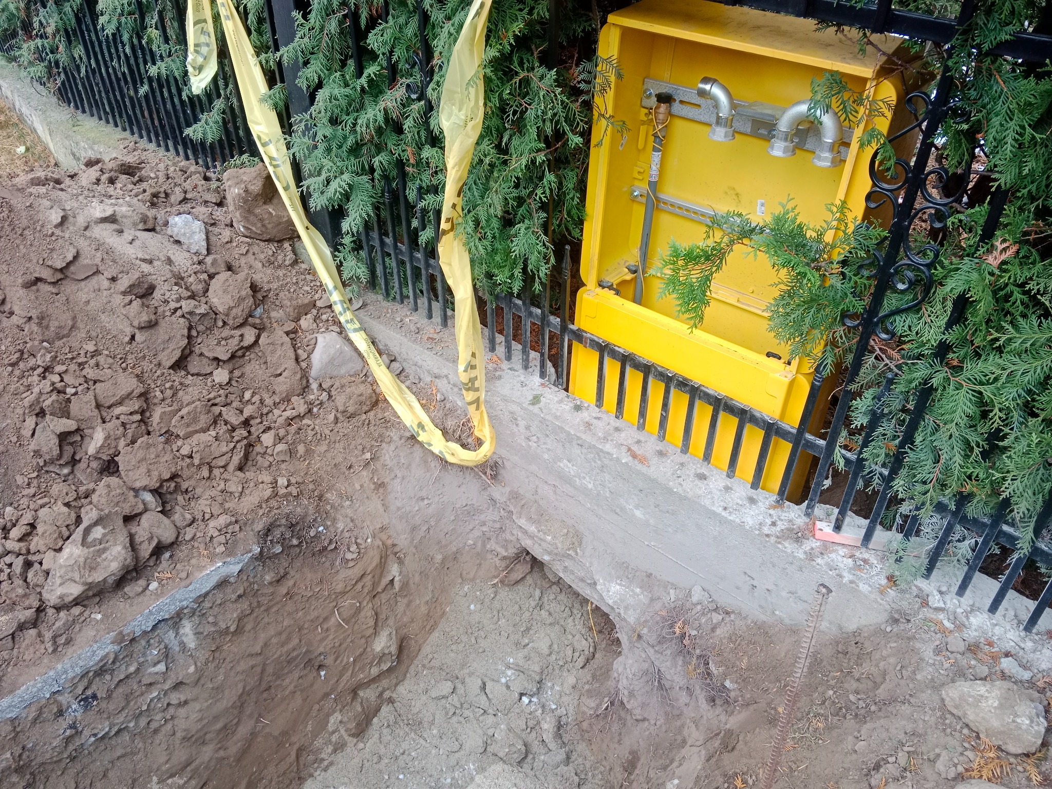 Wykop w ziemi z widocznym żółtym przyłączem gazowym osłoniętym metalowym płotkiem i taśmą ostrzegawczą. Prace ziemne w zielonym otoczeniu.