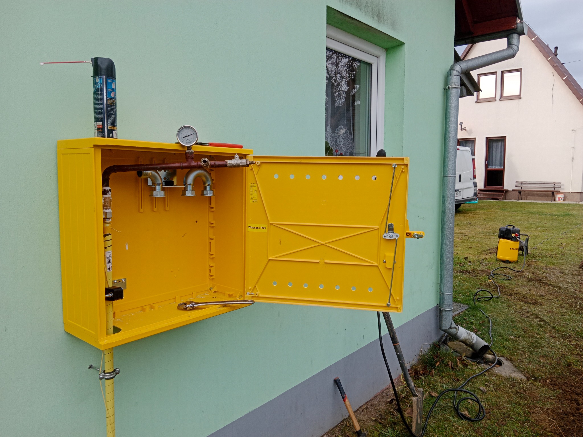 Żółta skrzynka gazowa zamontowana na ścianie budynku z otwartymi drzwiczkami, widoczne rury i manometr. Na górze puszka ze smarem, na dole klucz.