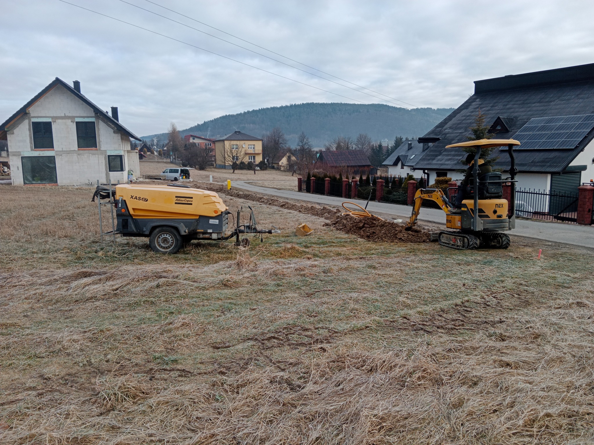 Mini koparka i kompresor Atlas Copco na działce podczas prac ziemnych. W tle domy i wzgórza.