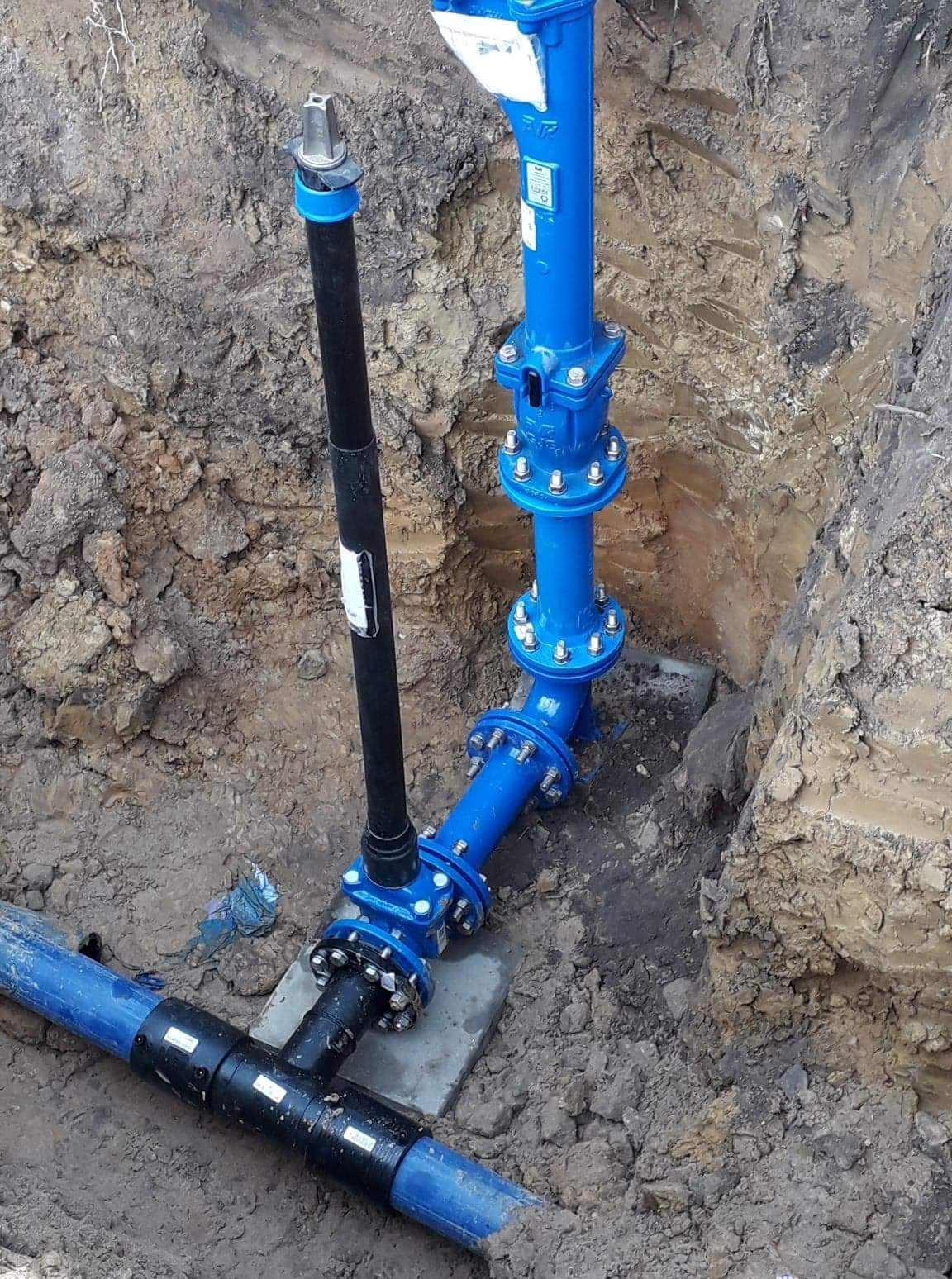 Niebieska instalacja rurociągowa z czarnym pionowym elementem, umieszczona w wykopie ziemnym. Widoczne złącza śrubowe i betonowe podpory.