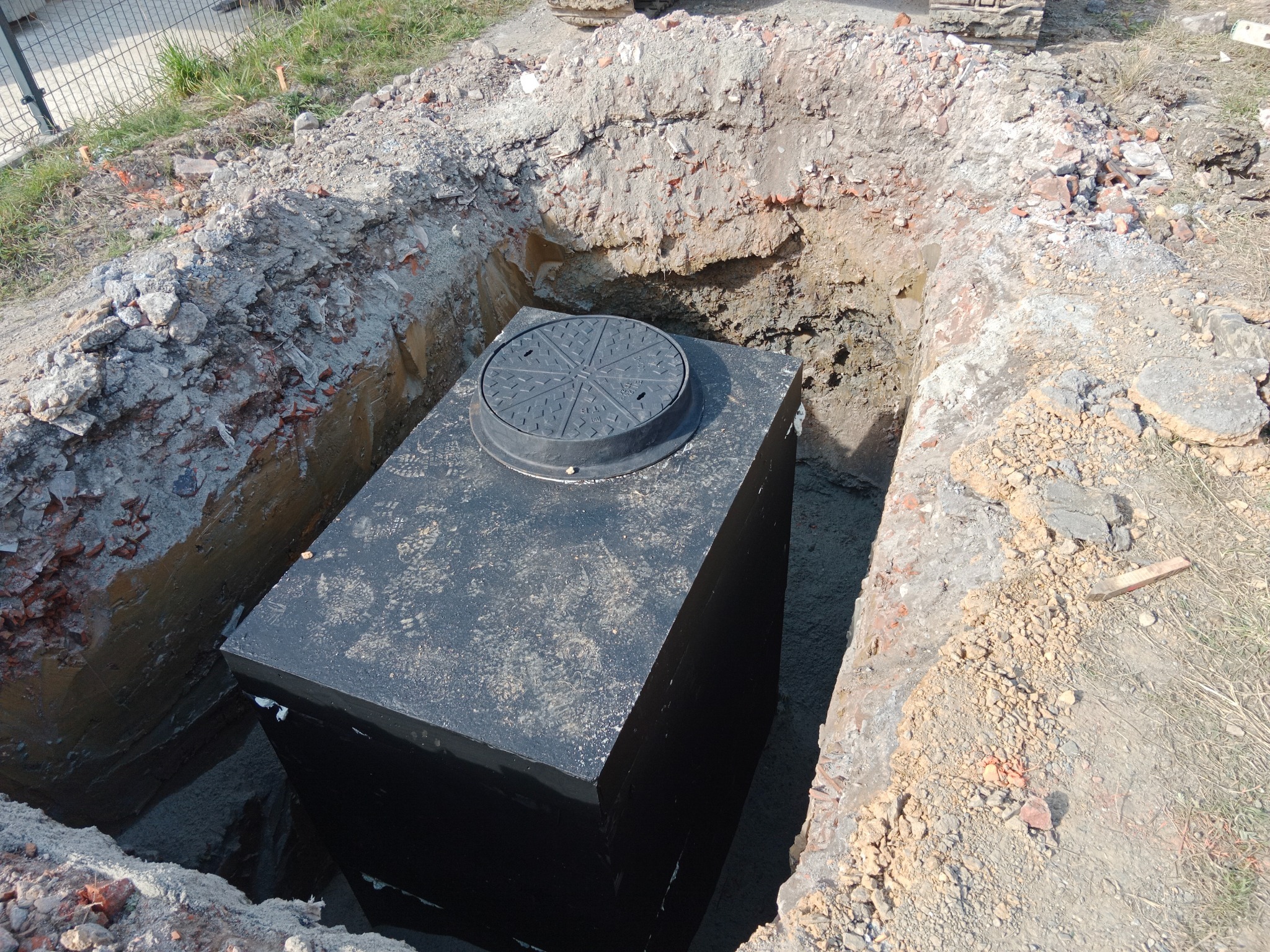 Czarny zbiornik z pokrywą w wykopie ziemnym, instalowany w Limanowej. Widoczne ślady montażu i otoczenie budowy.