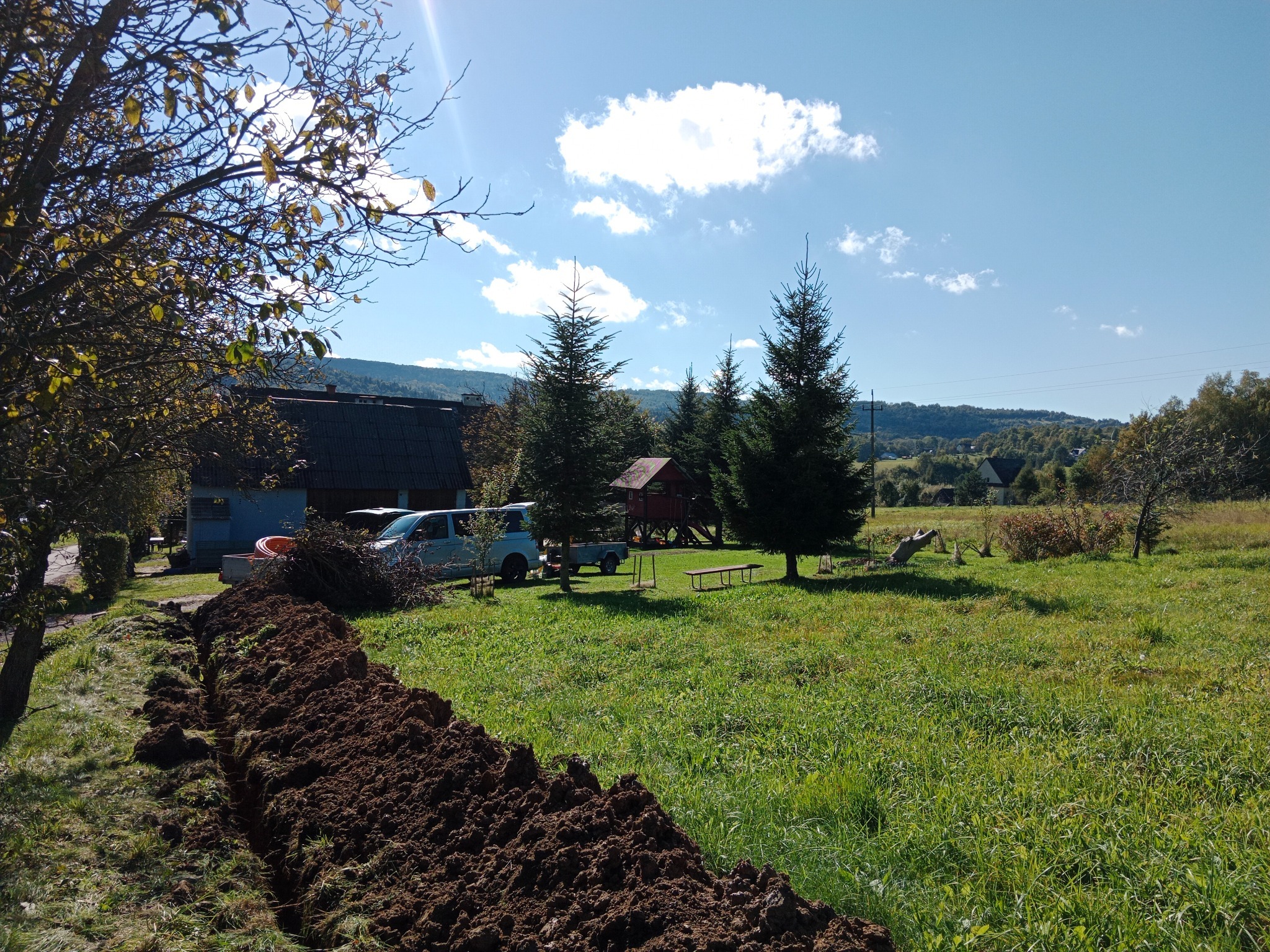 Wykonany wykop w ziemi, prawdopodobnie pod instalacje, na tle krajobrazu wiejskiego z domem i drzewami w słoneczny dzień. Widoczny samochód dostawczy.