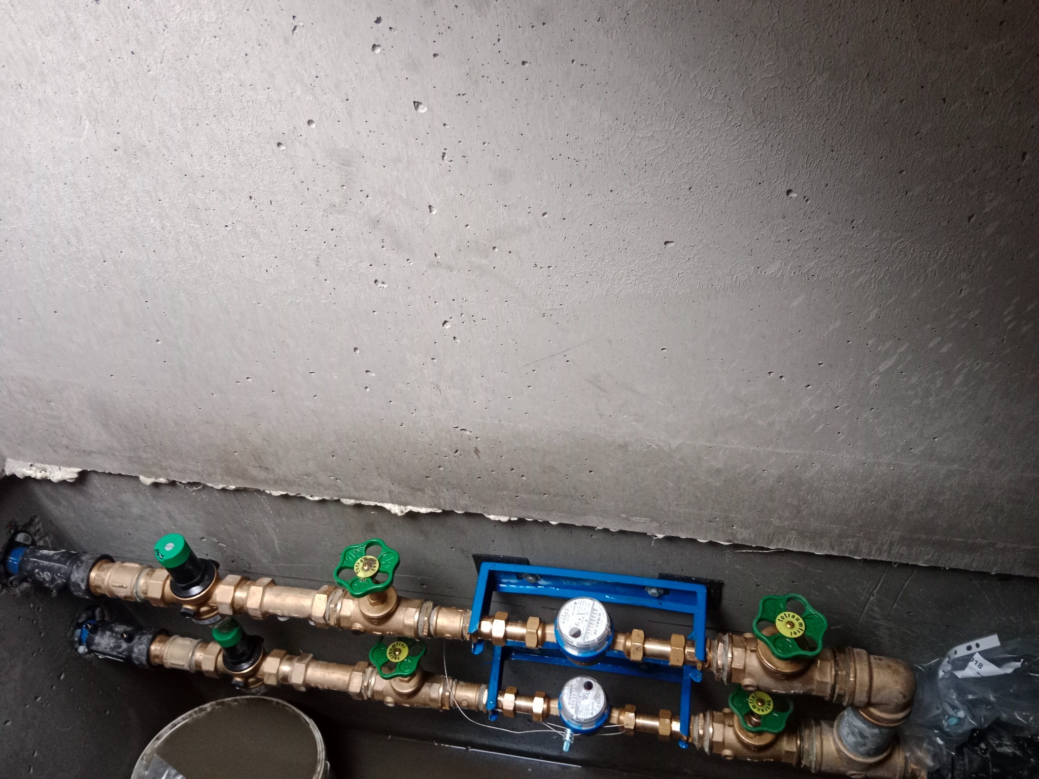 Dwa równoległe rzędy rur z zielonymi zaworami i niebieską ramą mocującą wodomierze na tle szarej, betonowej ściany. Instalacja wodna w piwnicy lub studzience.