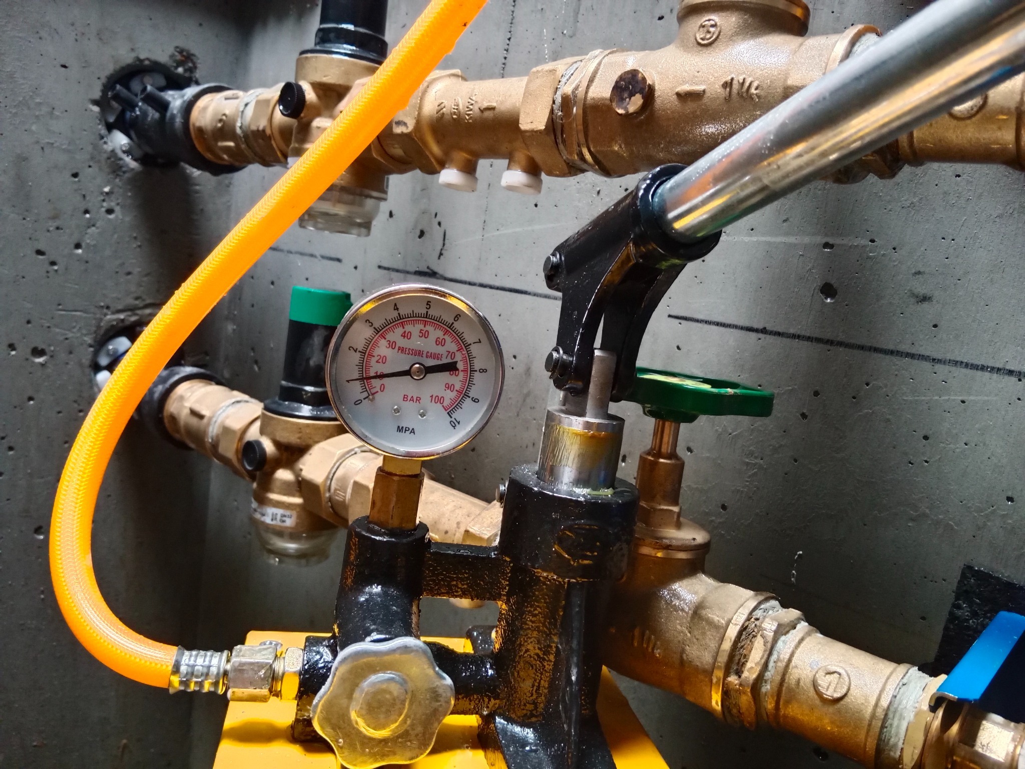 Instalacja wodna z manometrem i zaworami na tle surowego betonu. Widoczne mosiężne złączki i żółty przewód. Elementy instalacji wkomponowane w surowe, betonowe otoczenie.