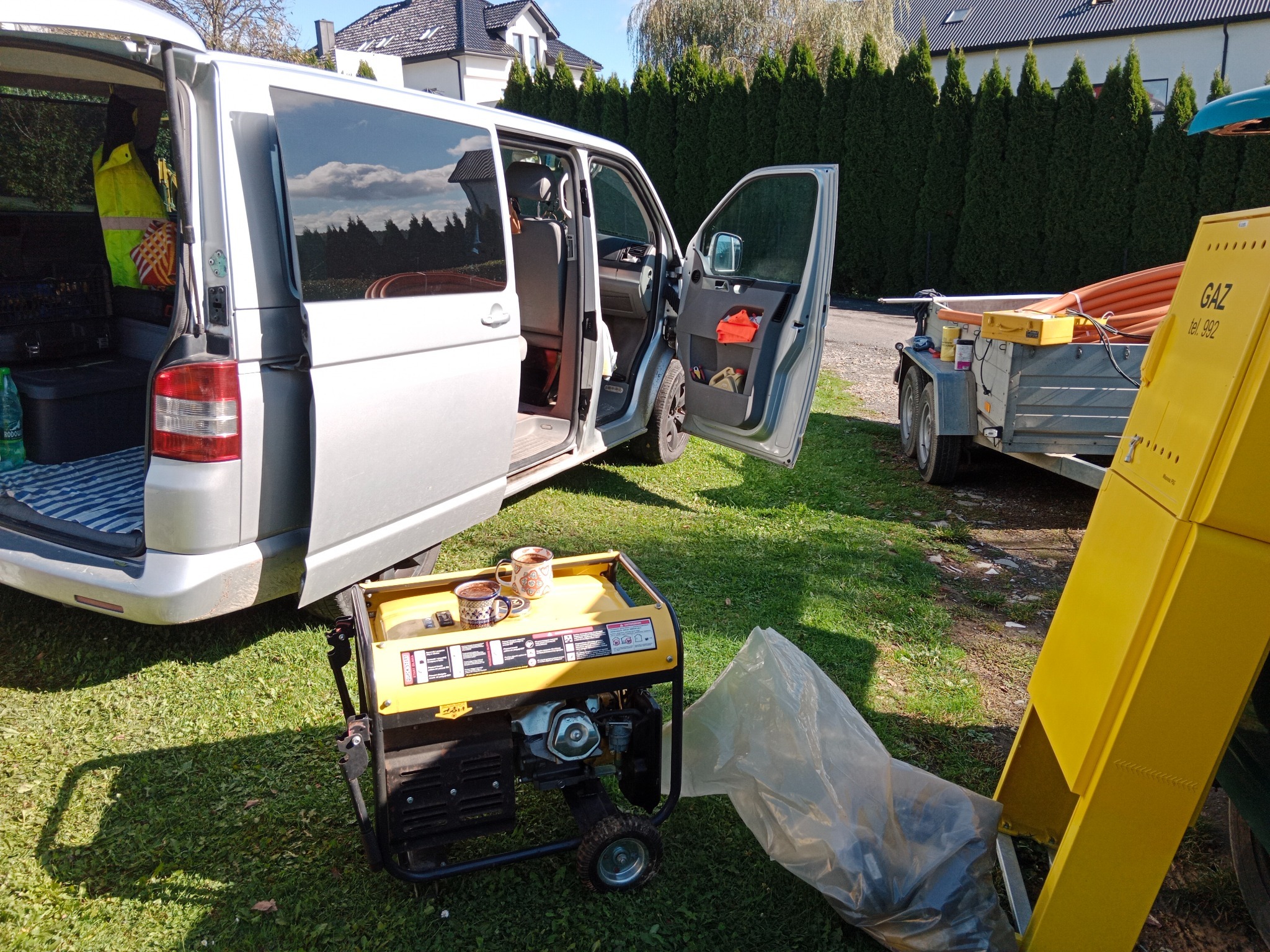 Sprzęt do prac ziemnych: agregat prądotwórczy z kubkami kawy, przyczepa z rurami, otwarty bus na trawie. Przerwa w pracy.