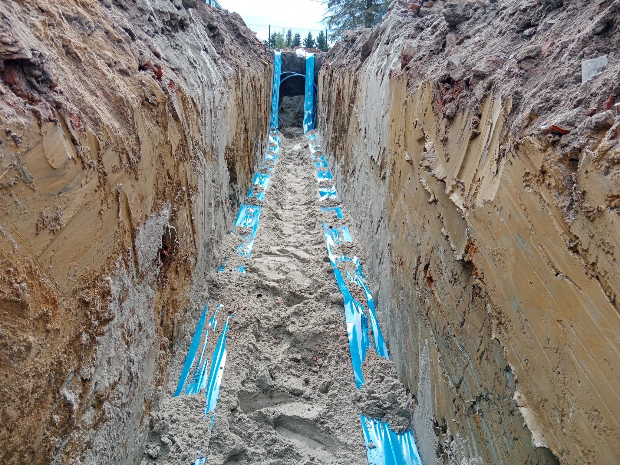 Wykop z ułożonymi rurami w niebieskiej folii ochronnej. Pionowe ściany wykopu z widoczną strukturą ziemi. Perspektywa z dołu wykopu.