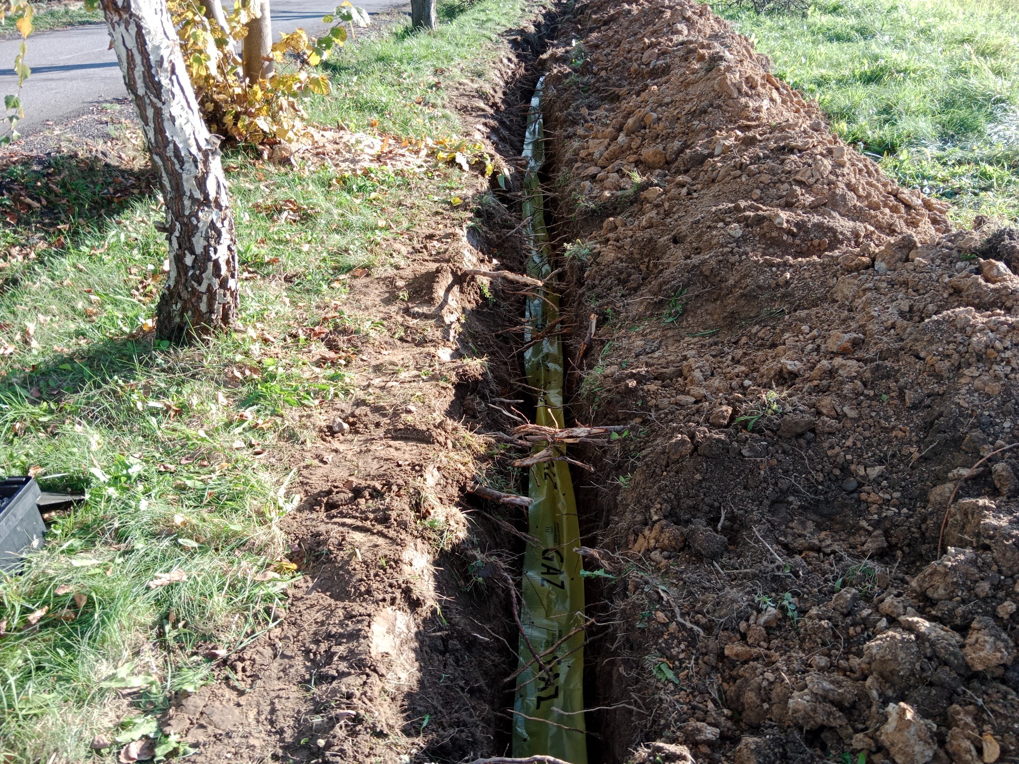 Wykop z ułożoną rurą gazową w zielonej osłonie, widoczny z góry. Po bokach ziemia i trawa. Rura z napisem 'GAZ'.