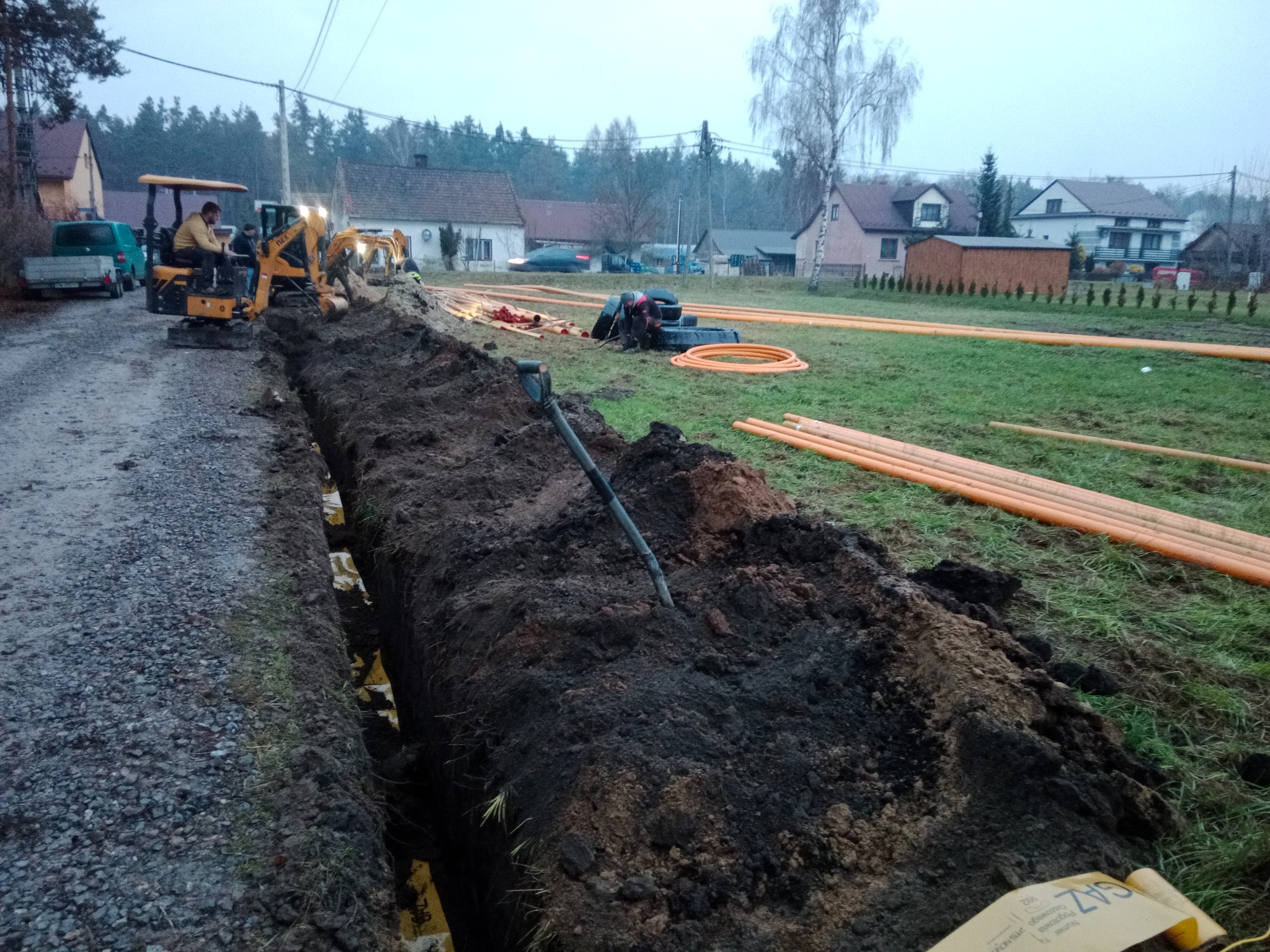 Prace ziemne: wykop pod rury, żółty mini-koparka w akcji, pomarańczowe rury ułożone na trawie. Domy w tle, pochmurne niebo.