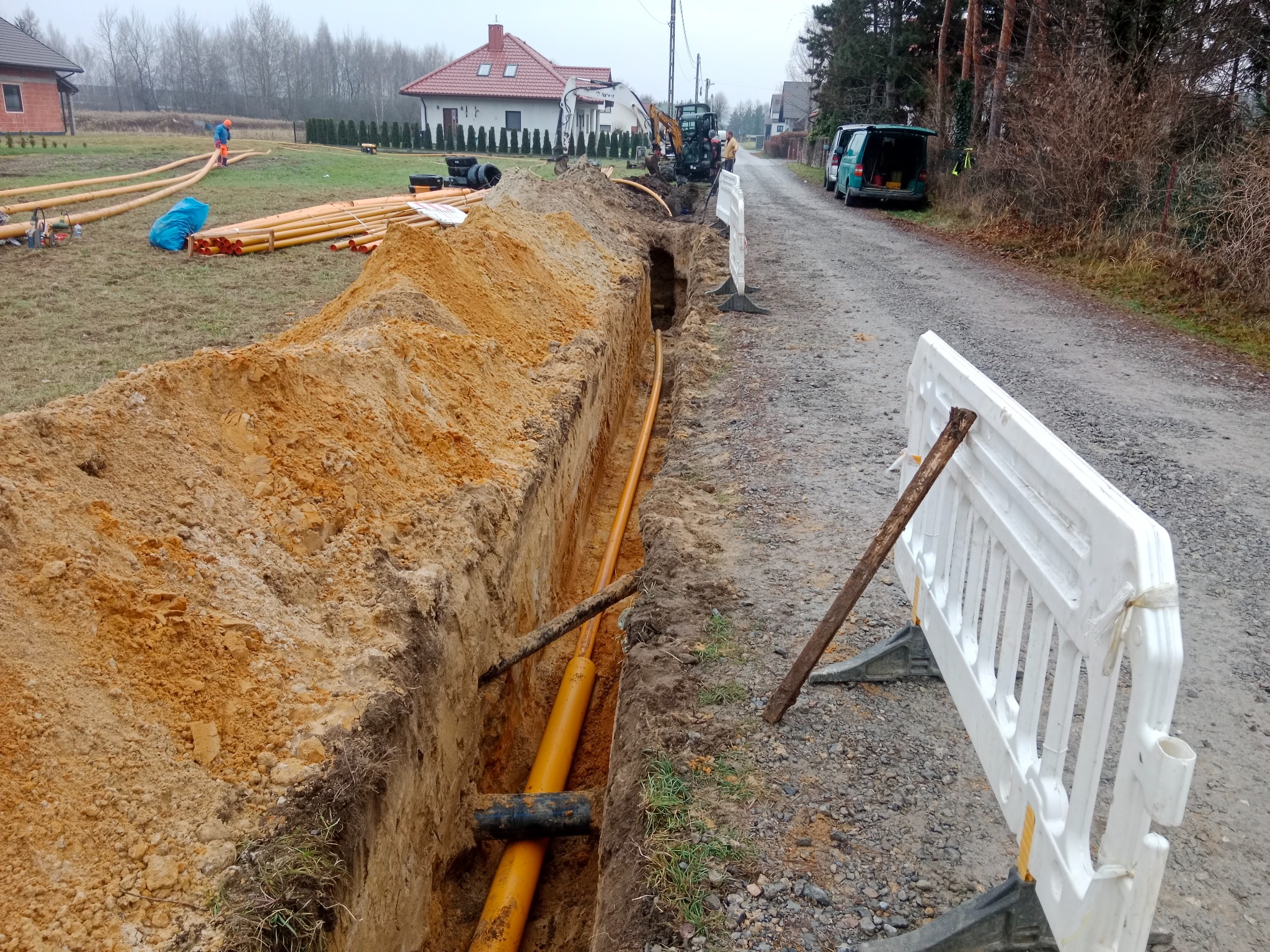 Żółte rury w wykopie ziemnym, zabezpieczone białą barierką. W tle domy jednorodzinne i sprzęt budowlany. Prace ziemne na etapie układania rur.